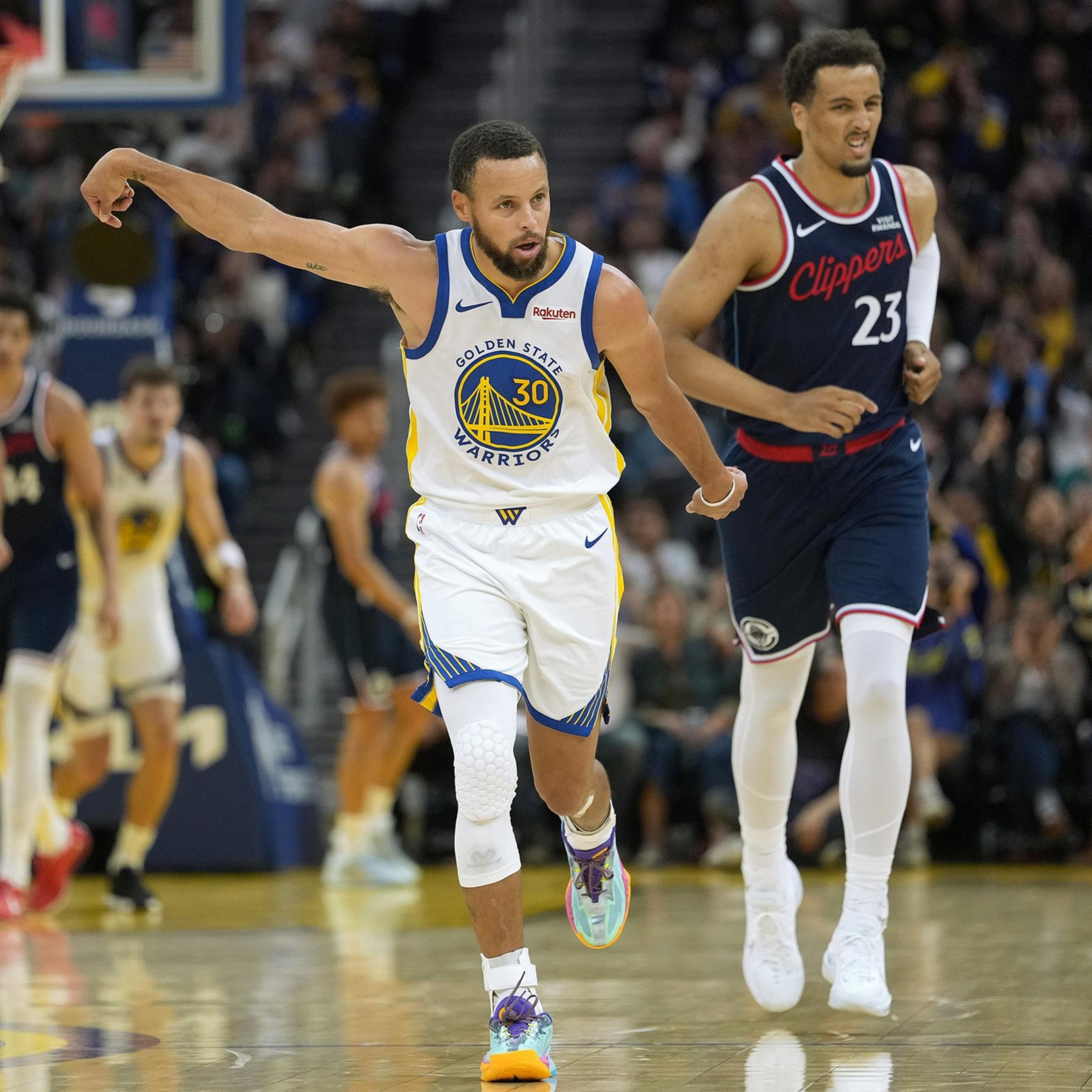 A Golden State Warriors player celebrates with an arm raised while a Clippers player runs behind him during a basketball game.