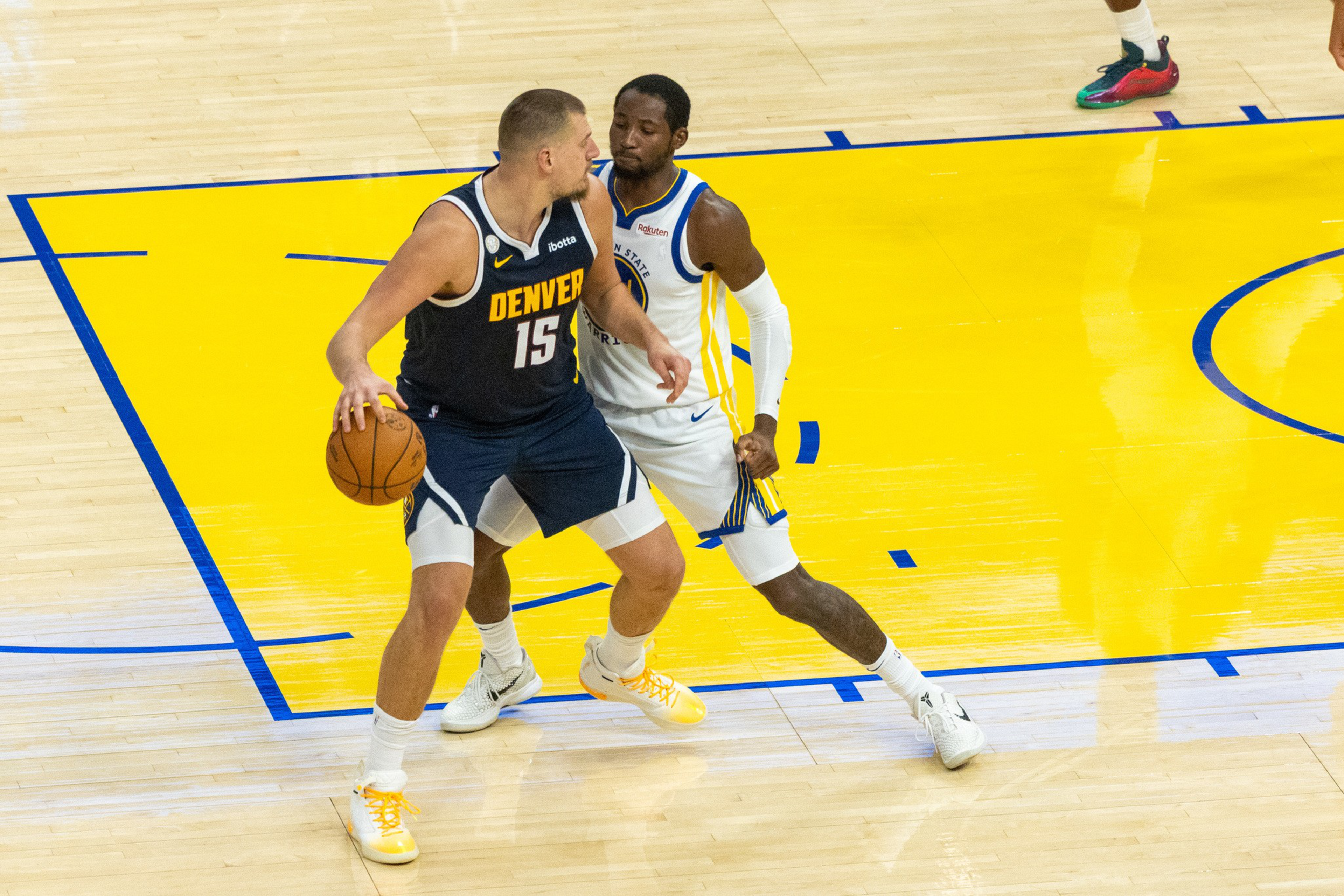 A Denver player with the ball is guarded closely by a Golden State player on a yellow and wood-colored basketball court.