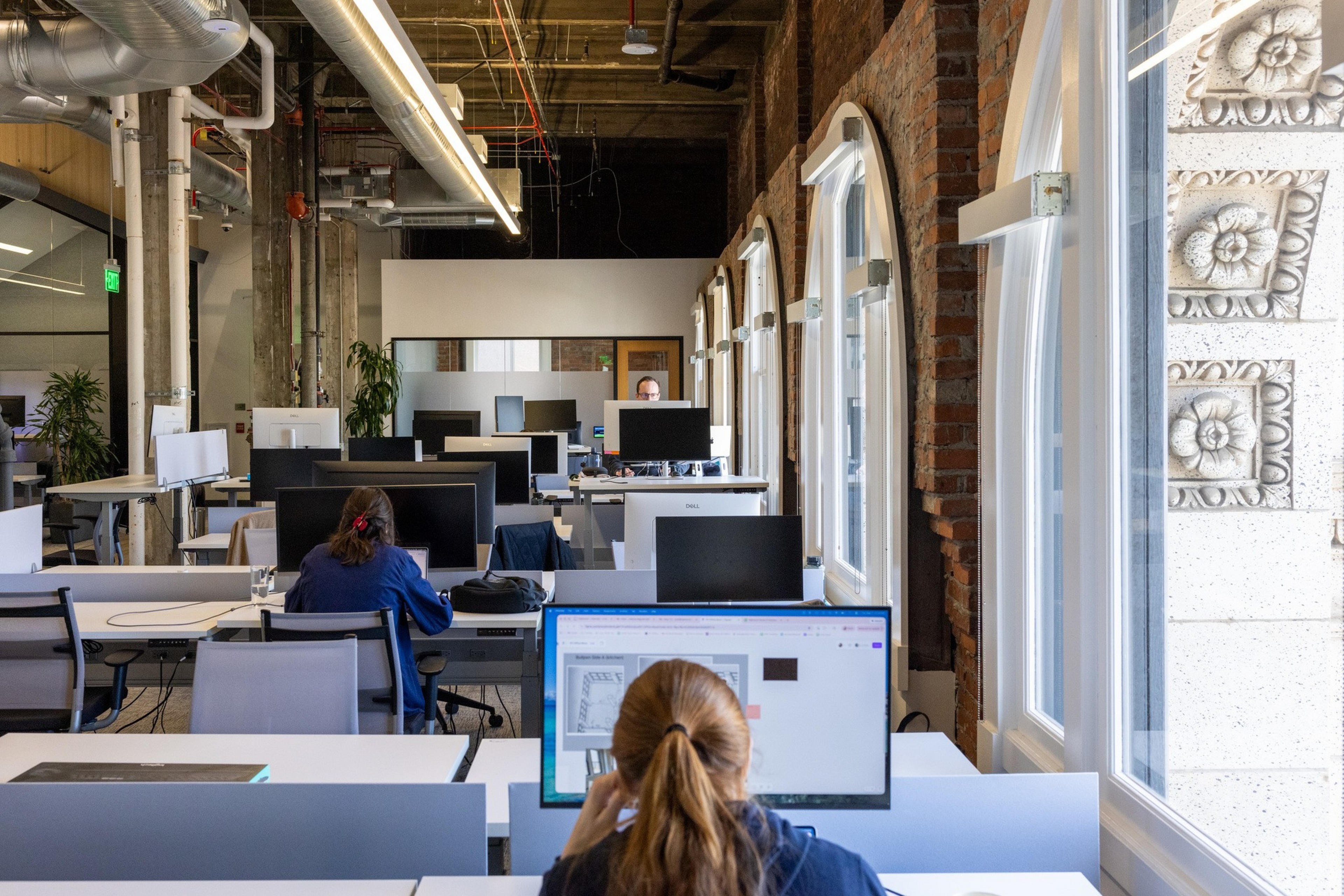 An open office space with exposed brick walls and large arched windows shows several people working at desks with computer monitors.