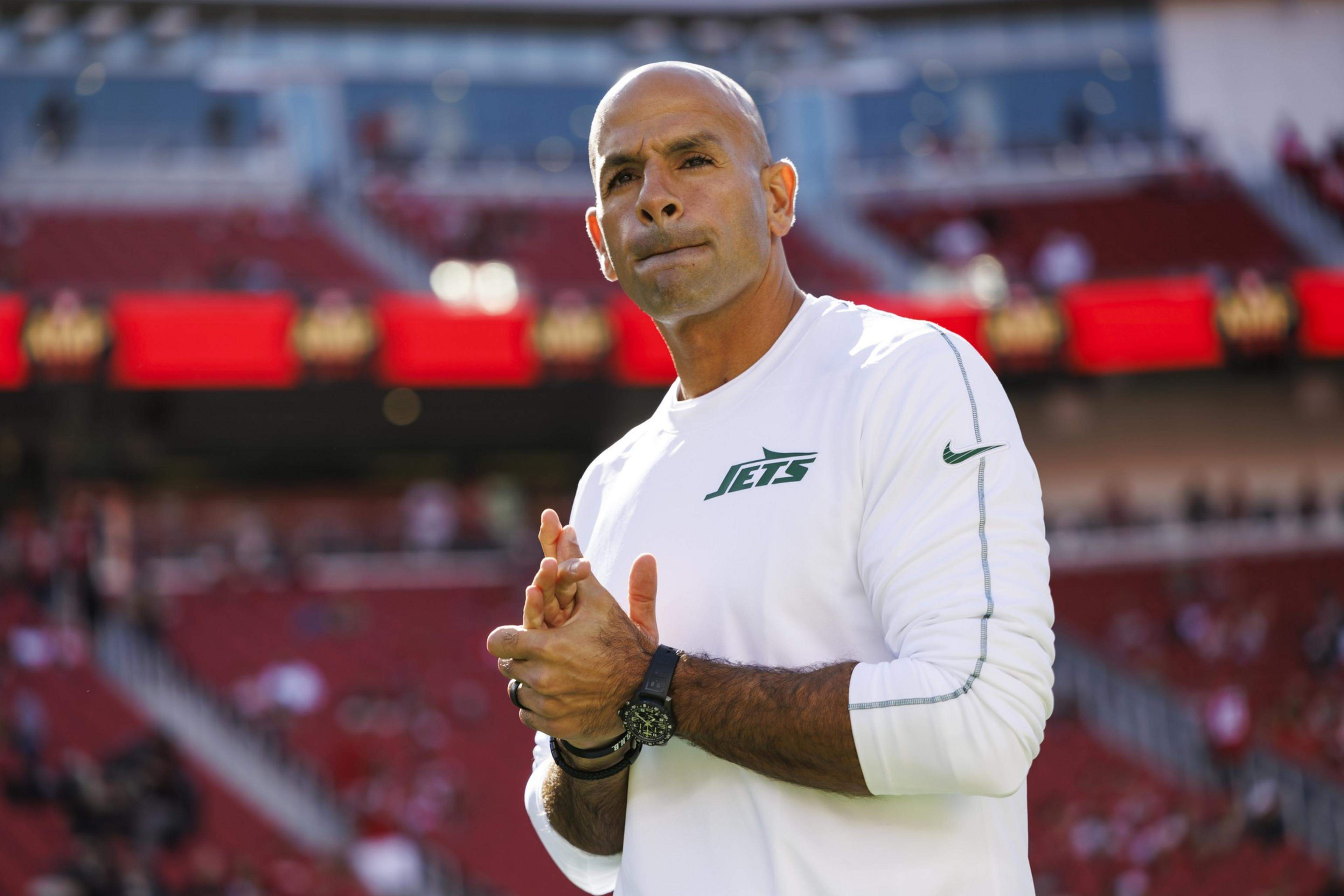 A person stands in a stadium wearing a white Jets shirt, with their hands clasped. The background shows blurred red stadium seats.