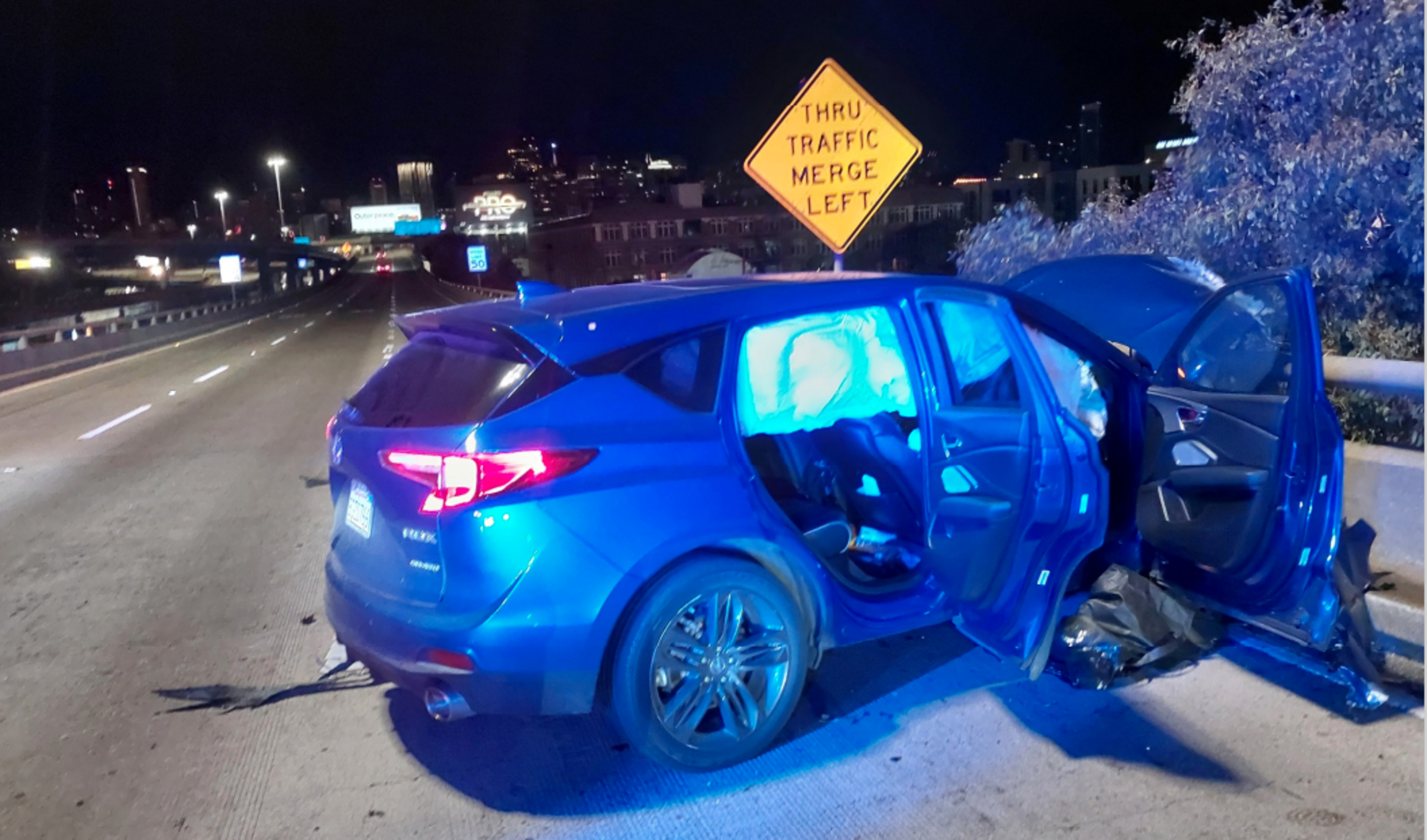 A blue car with deployed airbags and open doors is on a highway at night. Its front is damaged, and a &quot;Thru Traffic Merge Left&quot; sign is visible nearby.