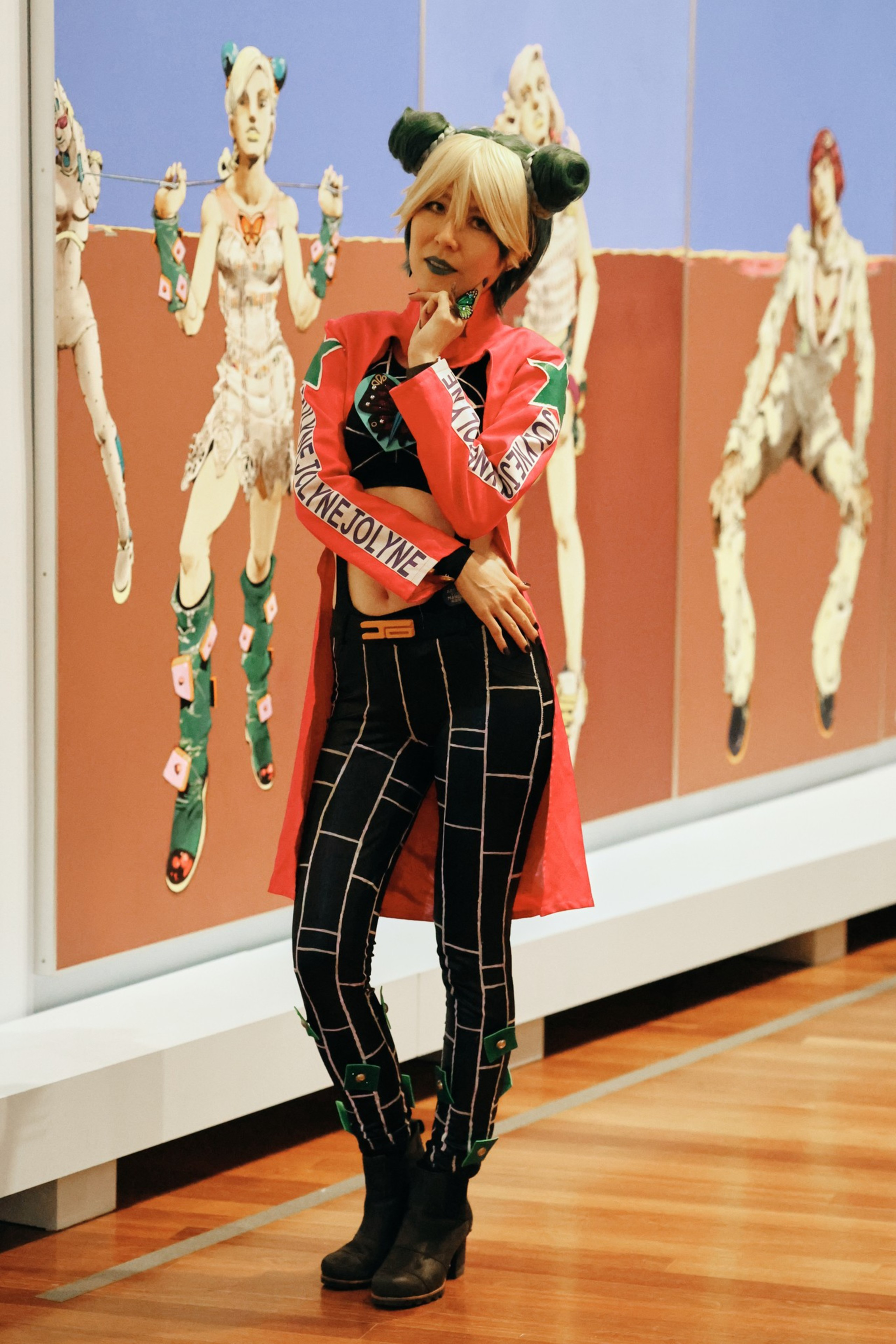 A person in a blonde wig with green buns and a red jacket poses in front of stylized artwork featuring figures in vibrant, detailed costumes.