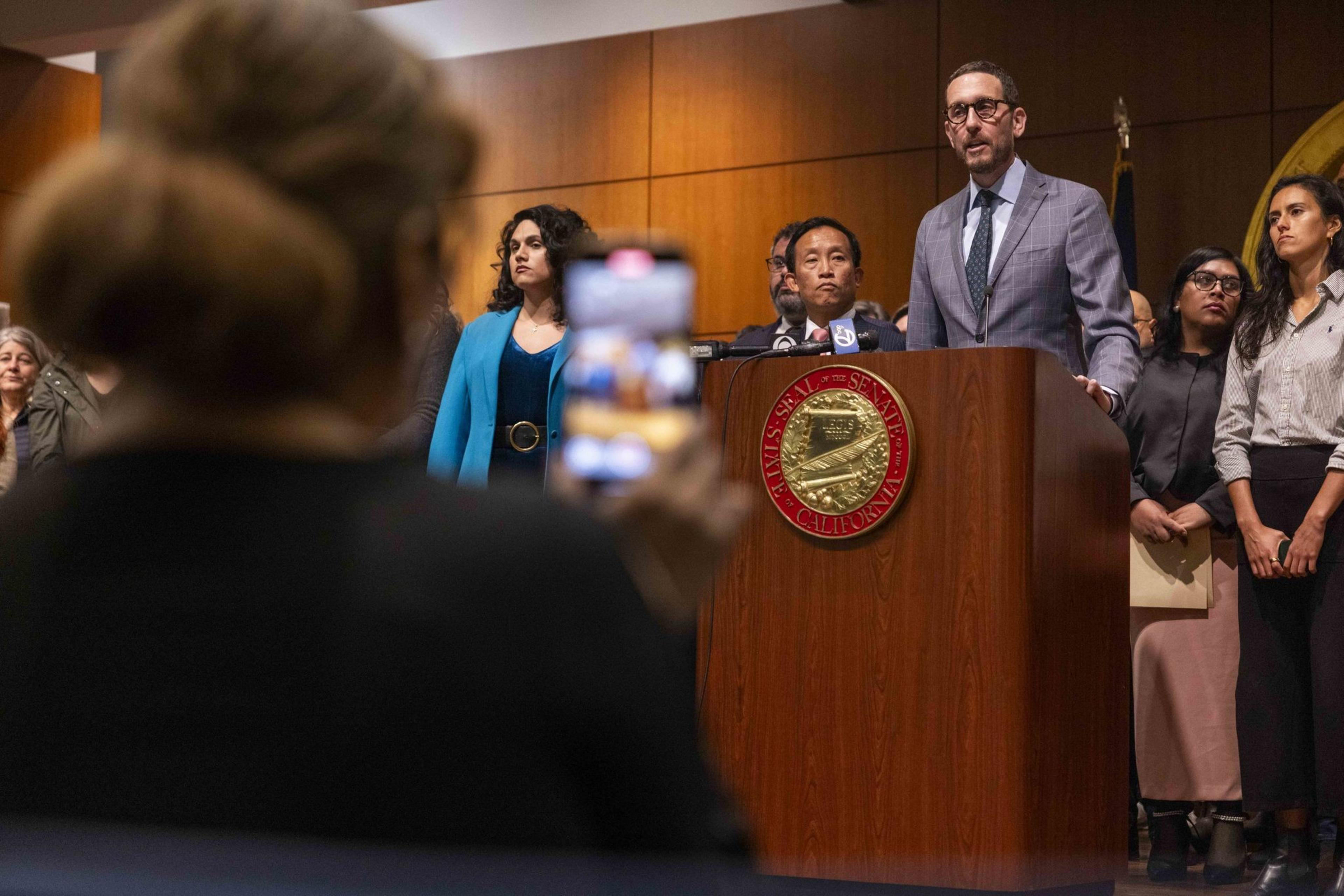 A group of people stand at a podium with microphones in a formal setting. A person in the foreground is taking a photo with a smartphone.