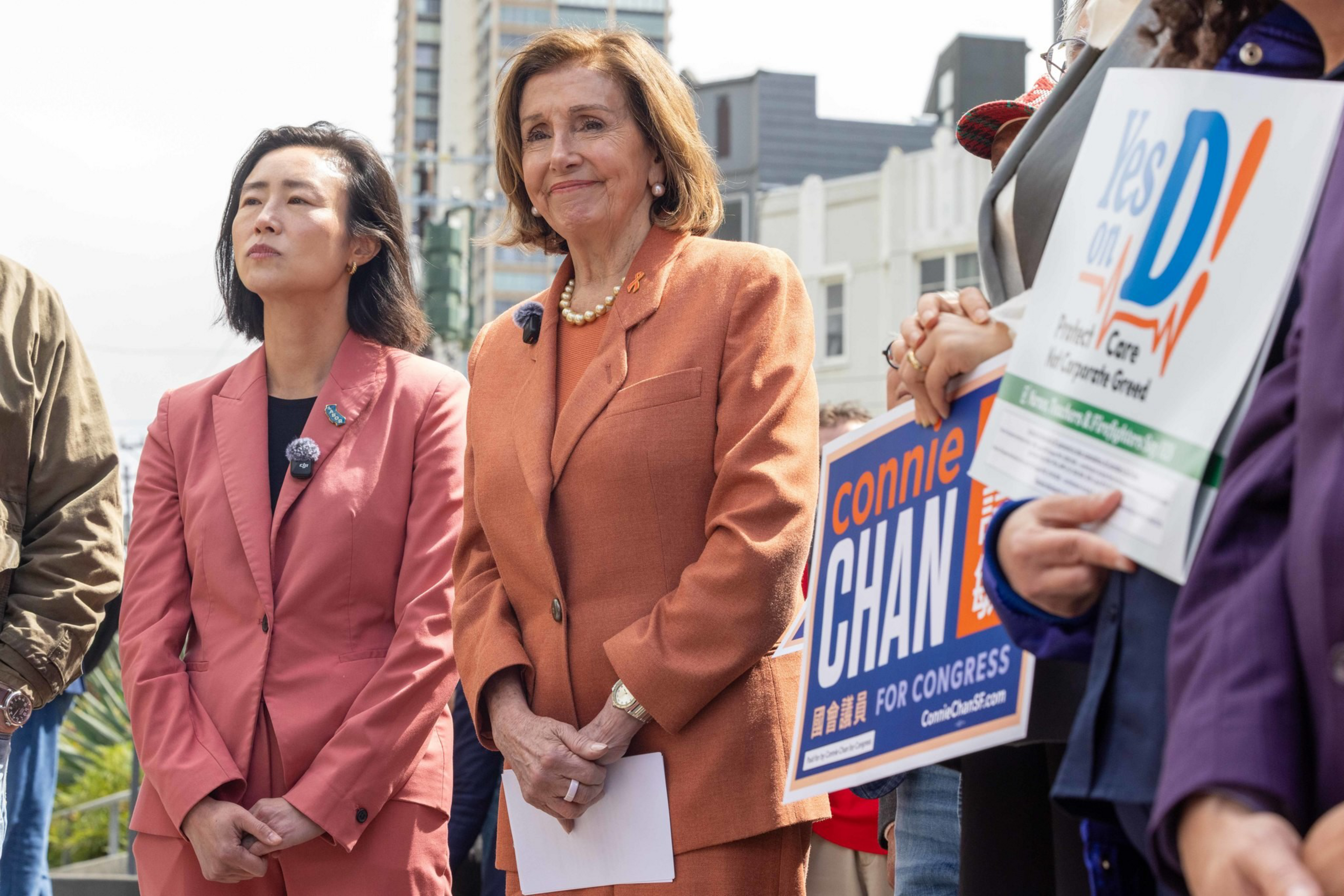 Two women stand side by side; one wears a coral suit and the other an orange suit, with campaign signs reading "Connie Chan for Congress" and "Yes on D" visible nearby.