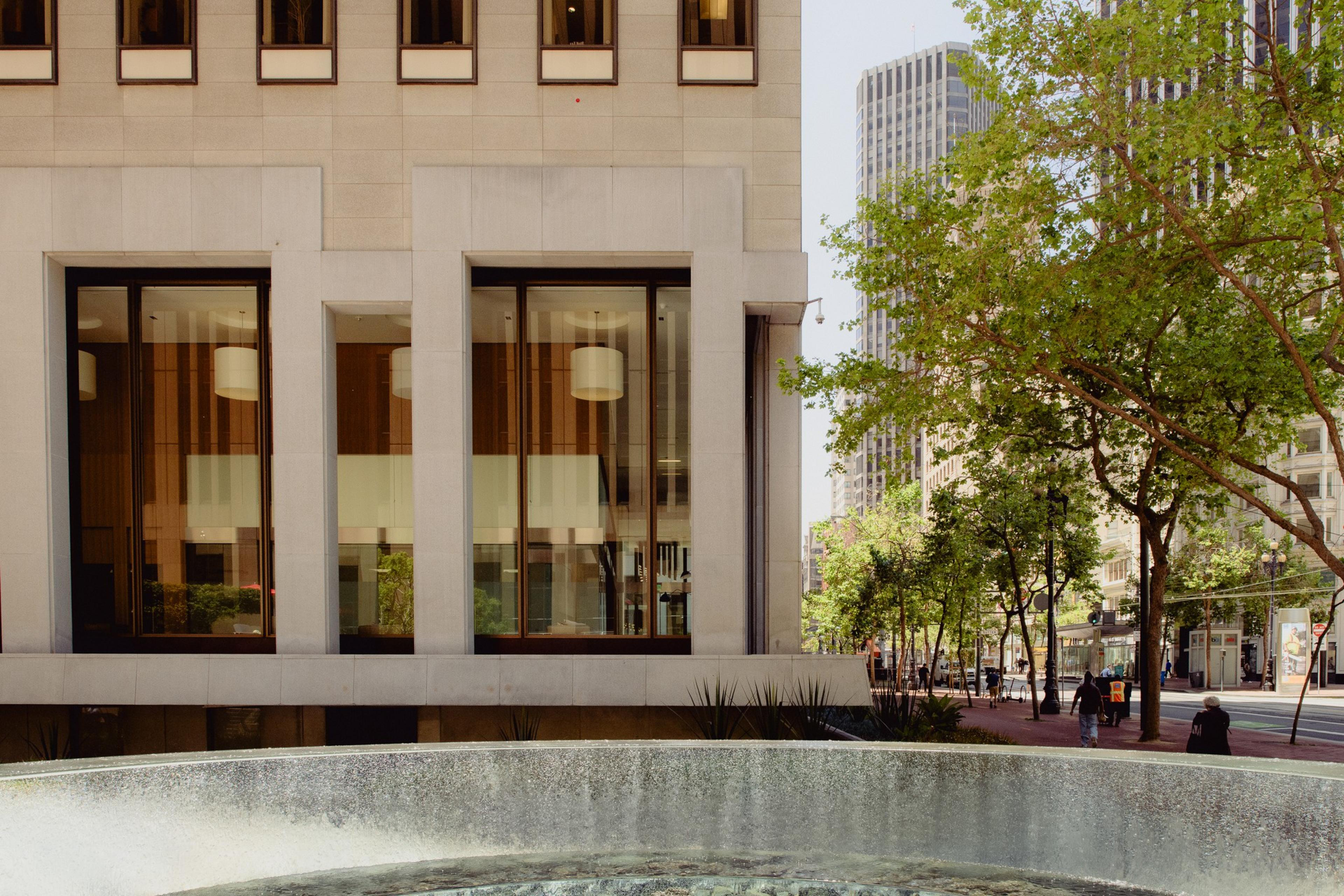 The image shows a modern building with large windows next to a tree-lined city street. In the foreground, there is a round water fountain.