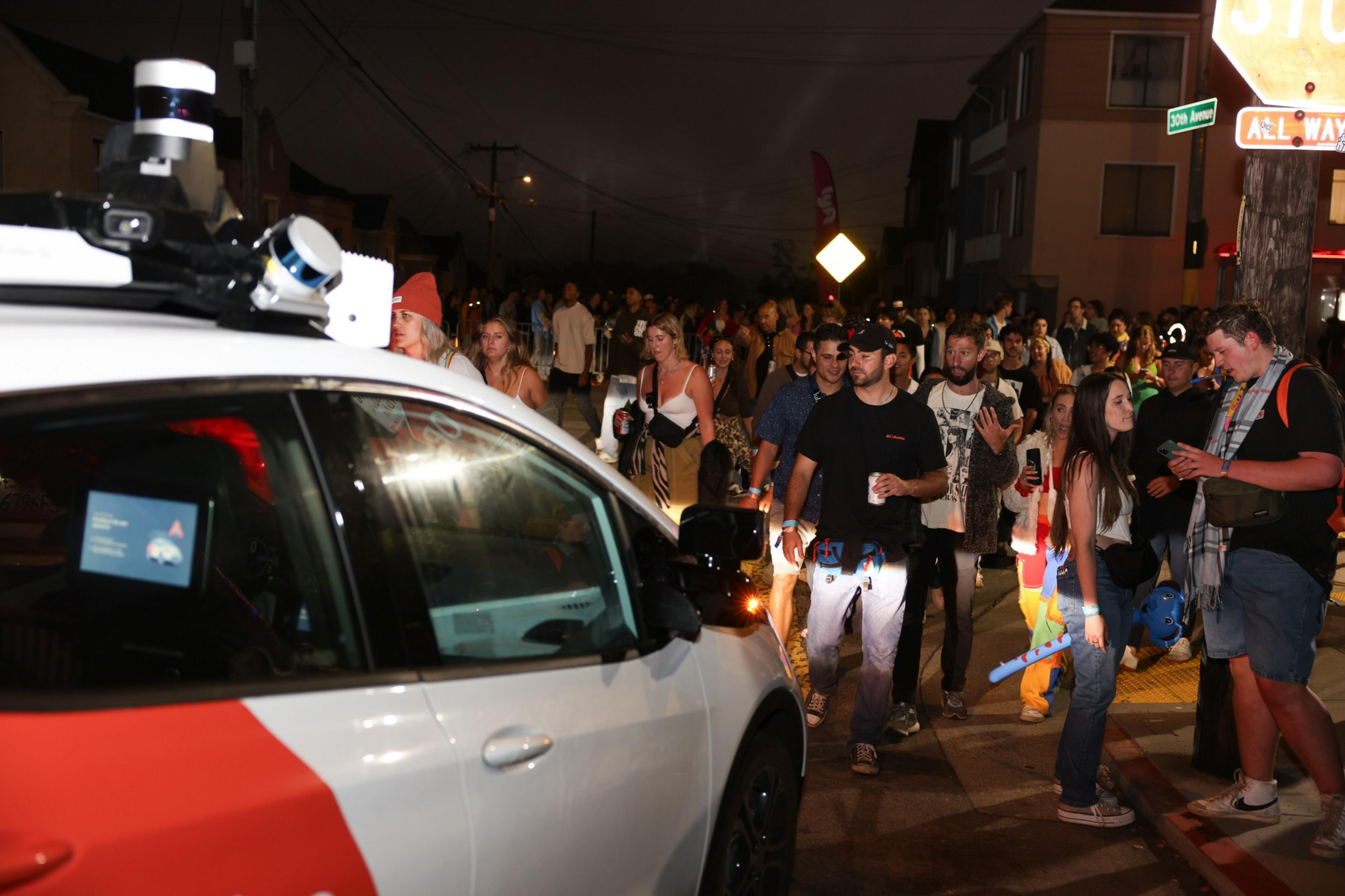 A large crowd of people walk along a street at night beside a white vehicle with mounted sensors under a stop sign.