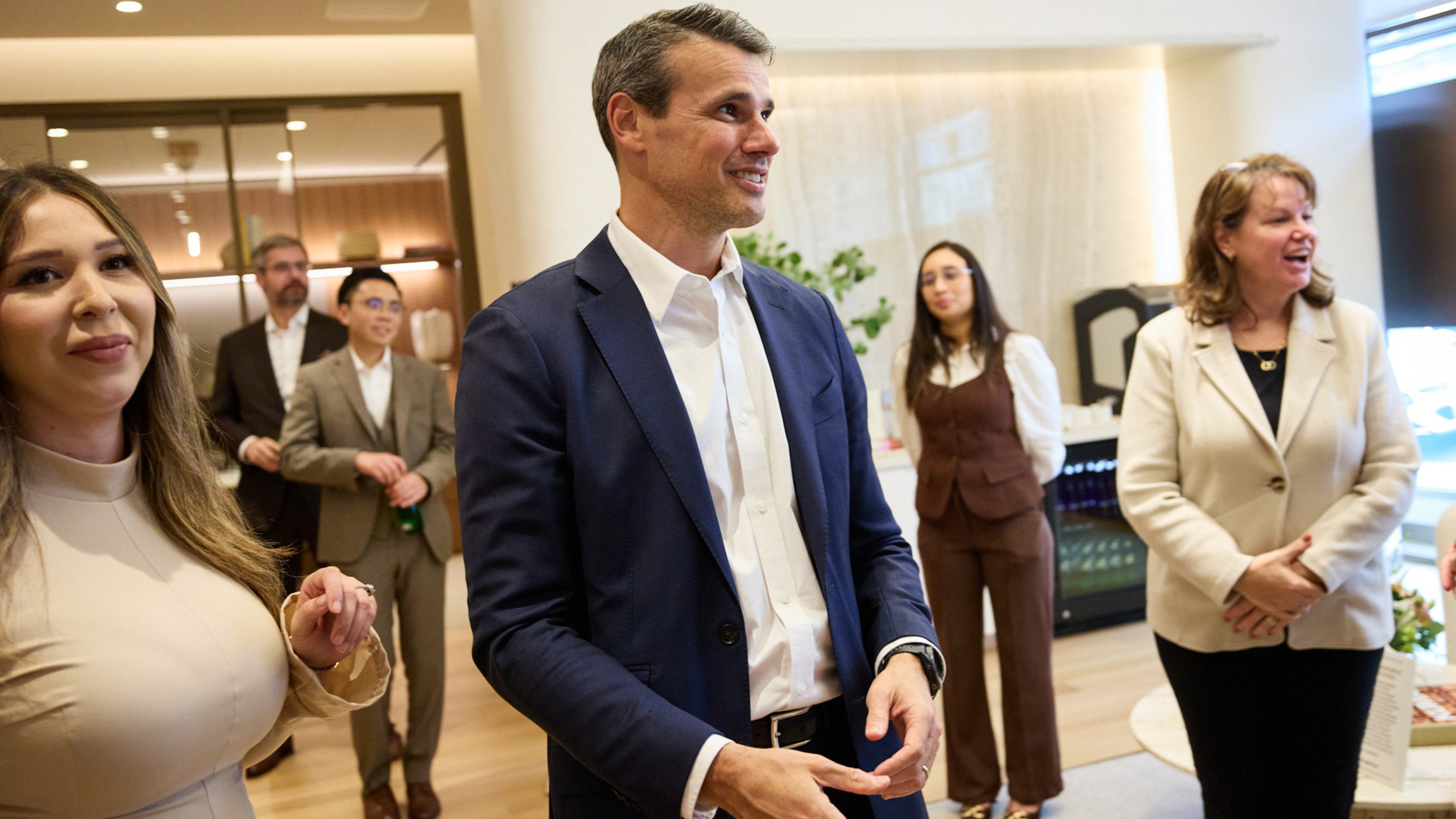 A group of six people, dressed in business attire, stand and smile in a modern office setting with light walls and wooden floors.