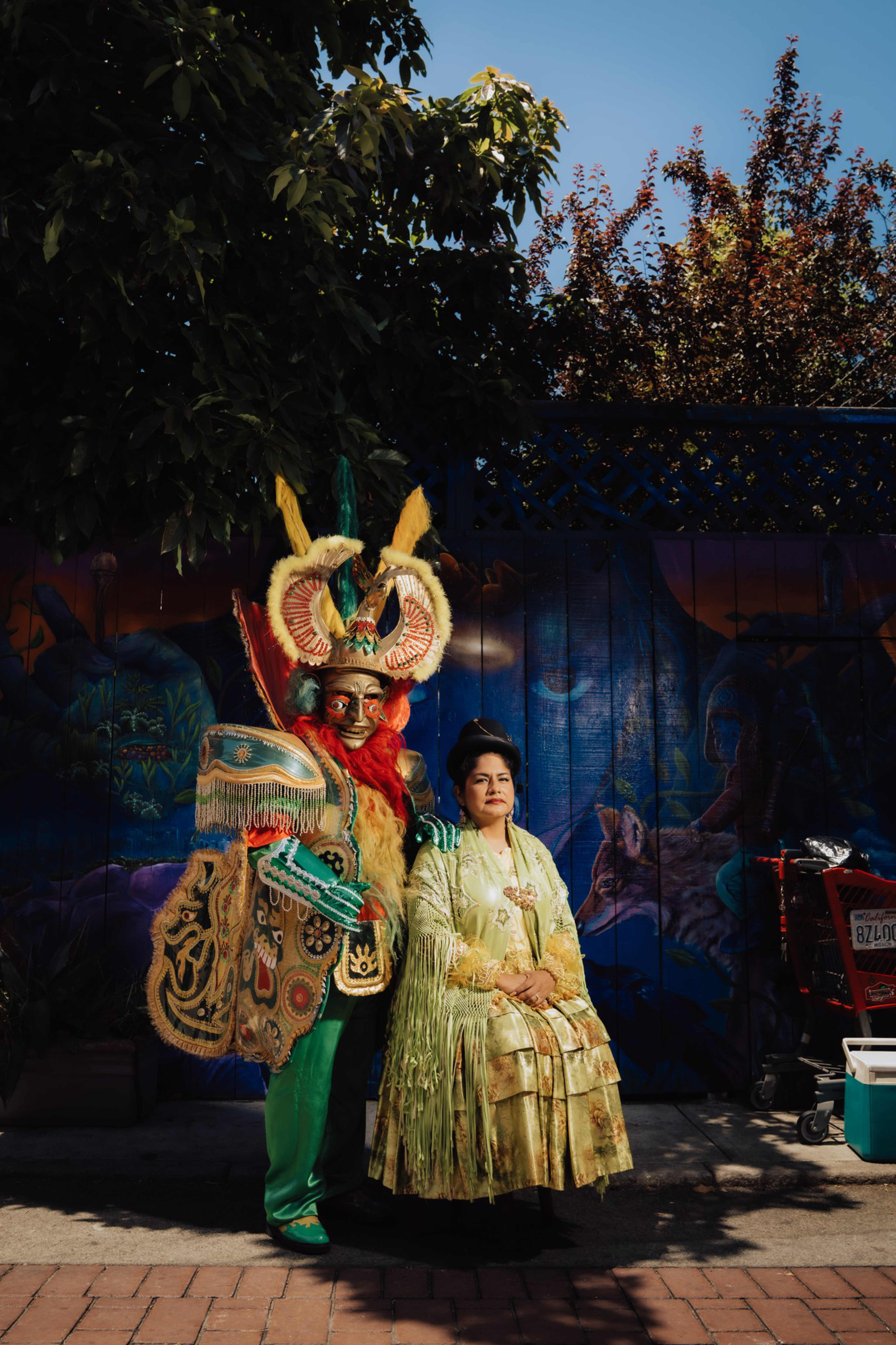 A person in an elaborate, colorful traditional costume stands beside a seated woman in a light green, fringed dress against a mural background.