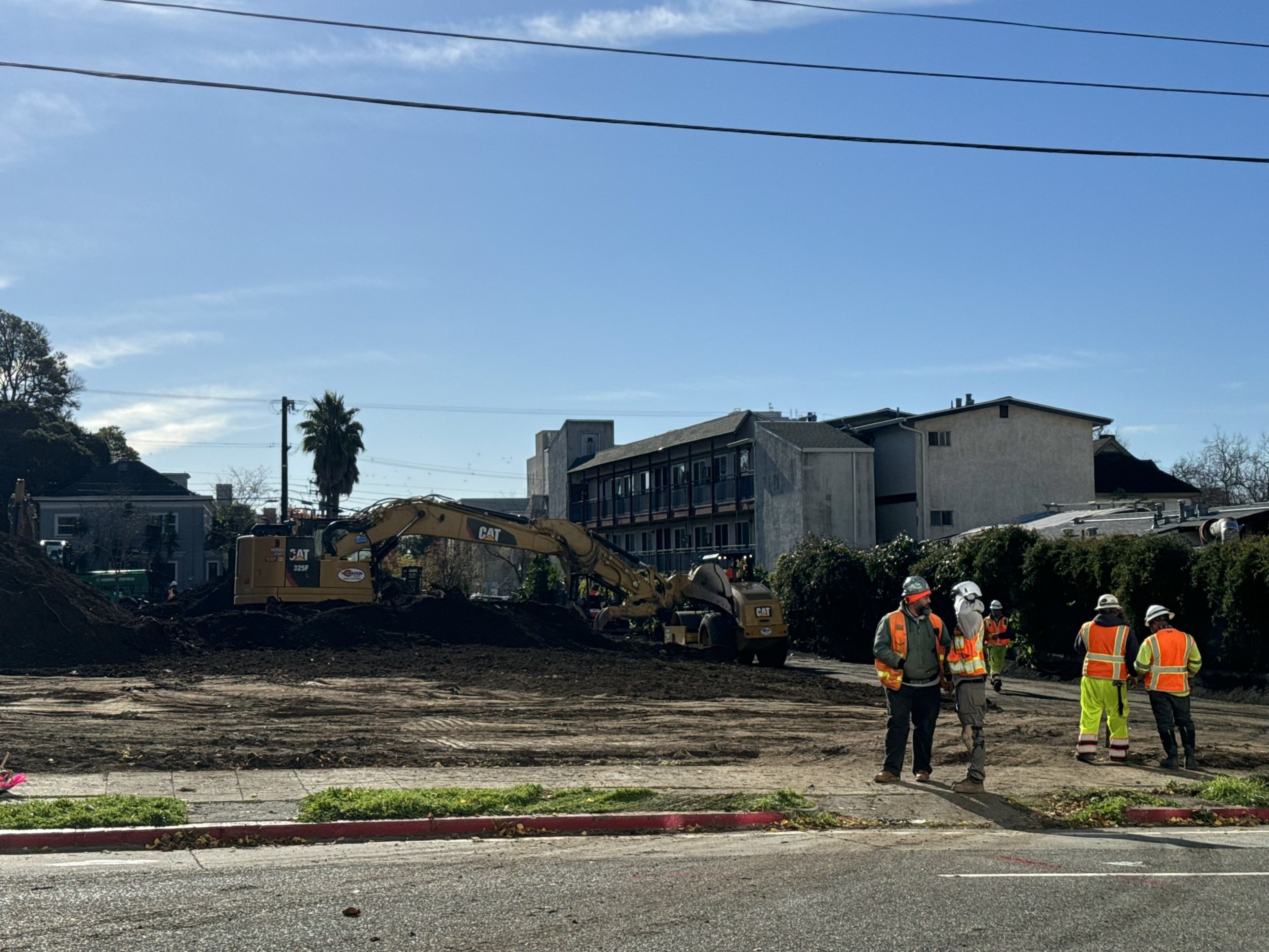 Construction workers stand to the left while a excavator digs up dirt on a plot of land.