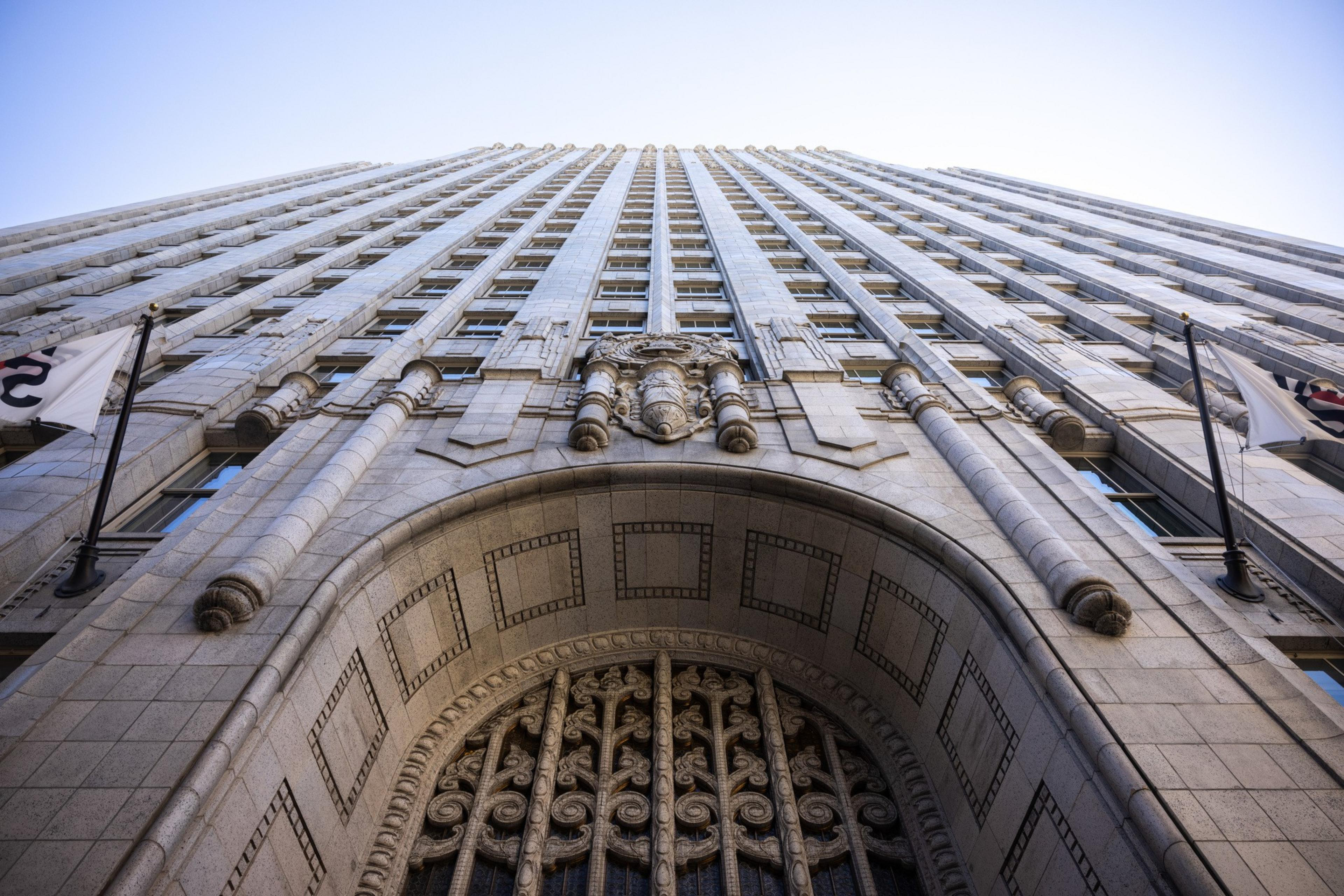 The image shows an upward view of a tall, ornate stone building with intricate architectural details, large windows, and flags on either side of the entrance.