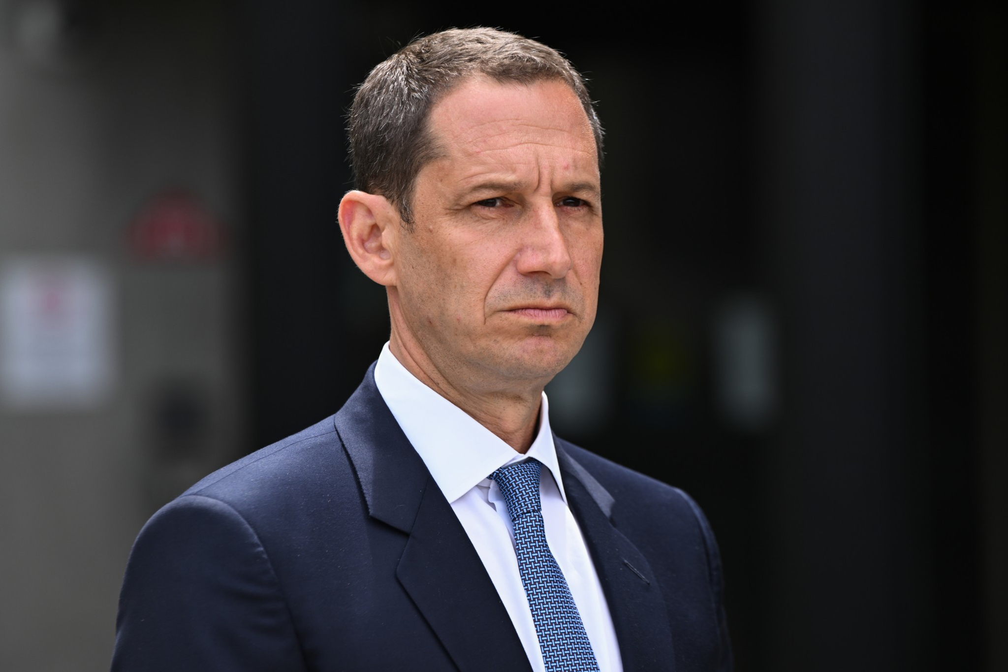 A man with short dark hair and a serious expression wears a dark suit, white shirt, and blue patterned tie against a blurred dark background.