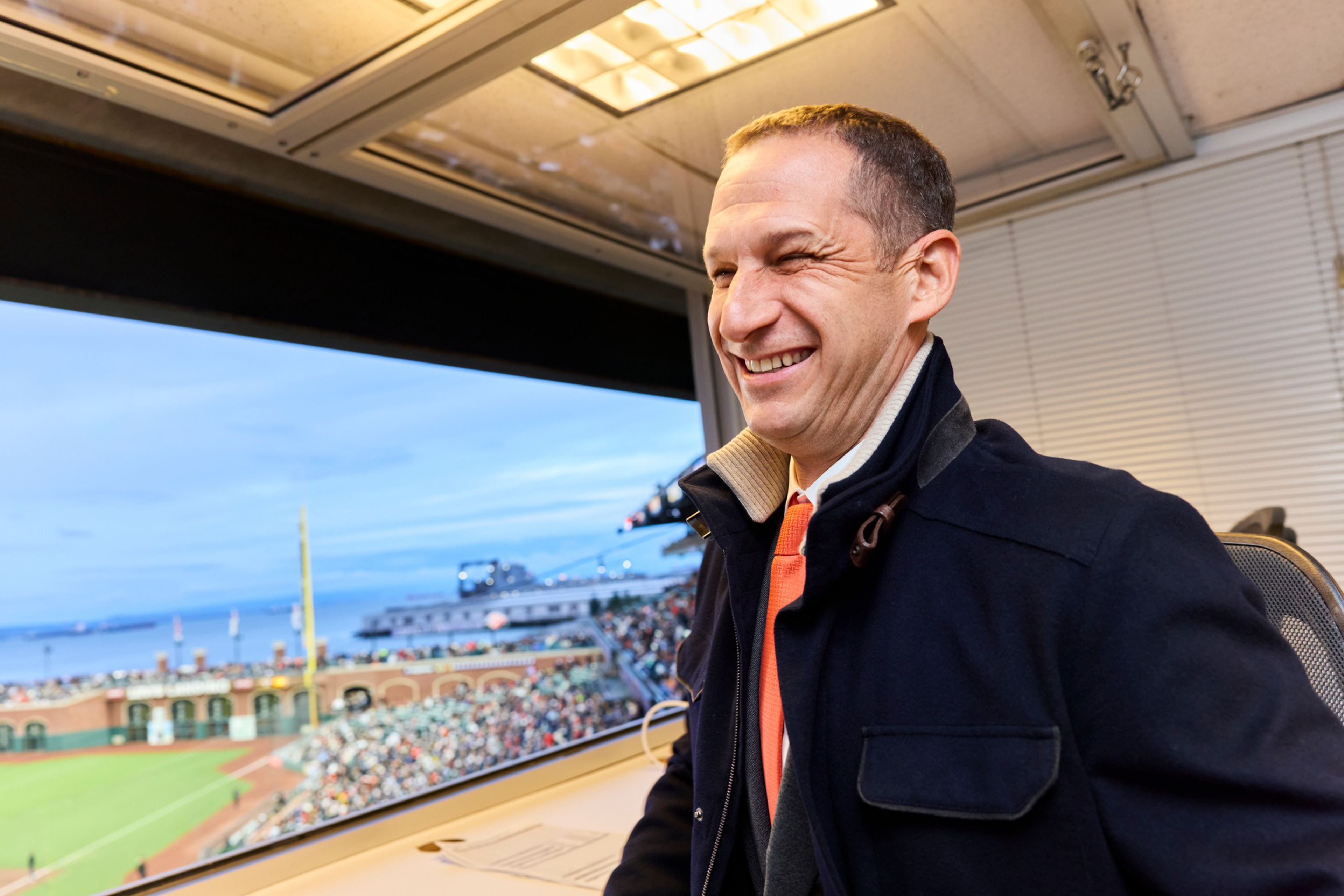 A smiling man in a dark coat and orange tie sits inside a sports press box overlooking a baseball stadium with a crowd at dusk.