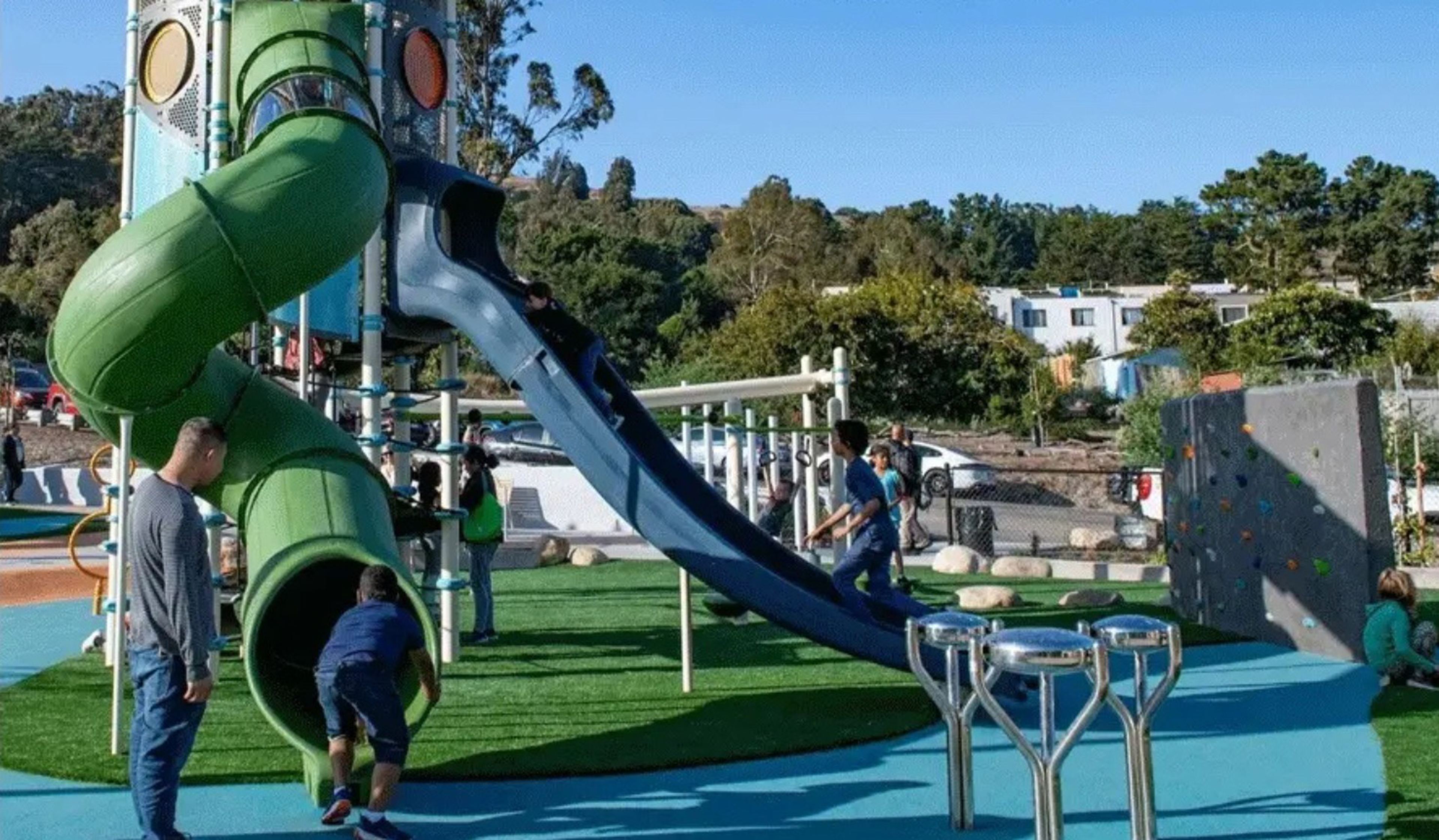 Kids play on a slide structure in a playground as an adult looks on. 