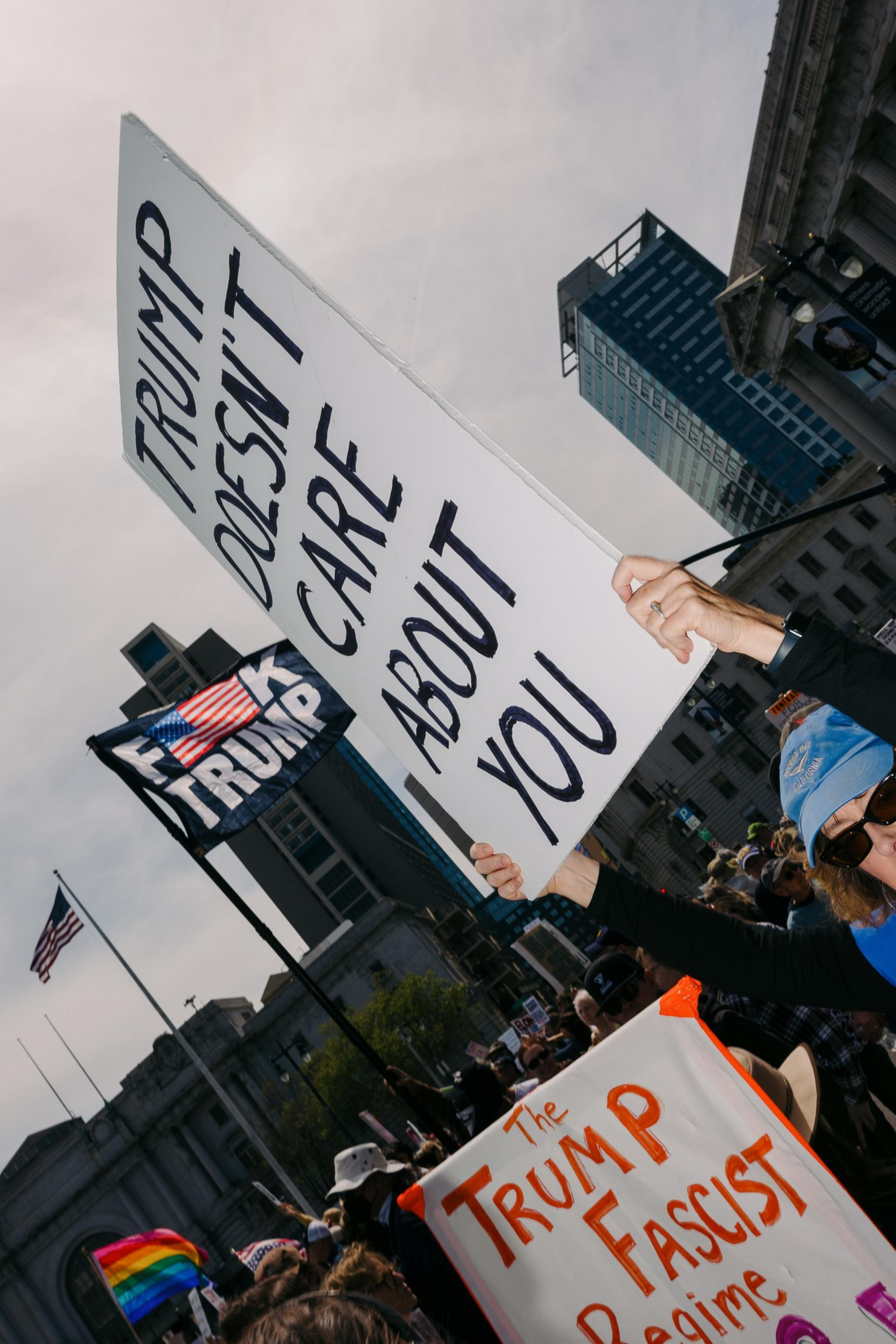 Protesters hold signs critical of Trump, with an American flag and buildings in the background. One sign says &quot;Trump doesn’t care about you.&quot;