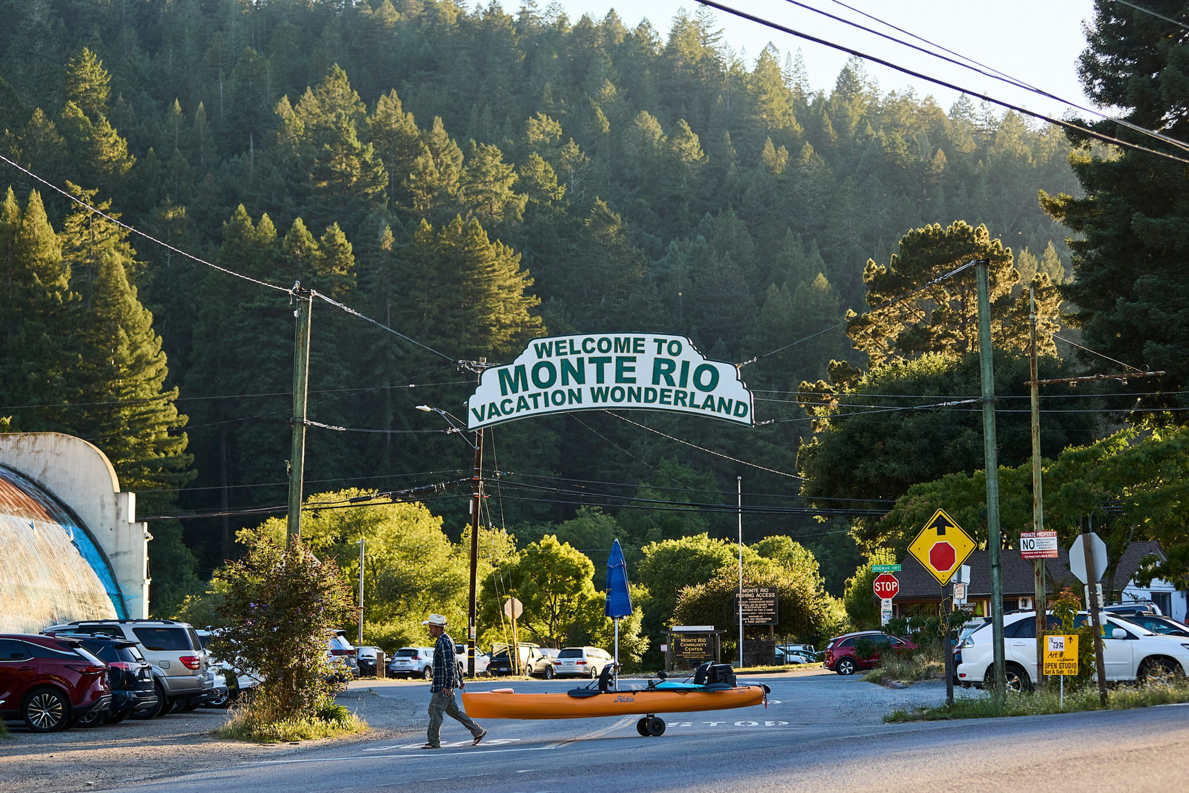 A person walks across a gravel road pulling an orange kayak on wheels beneath a sign that reads “Welcome to Monte Rio Vacation Wonderland.”