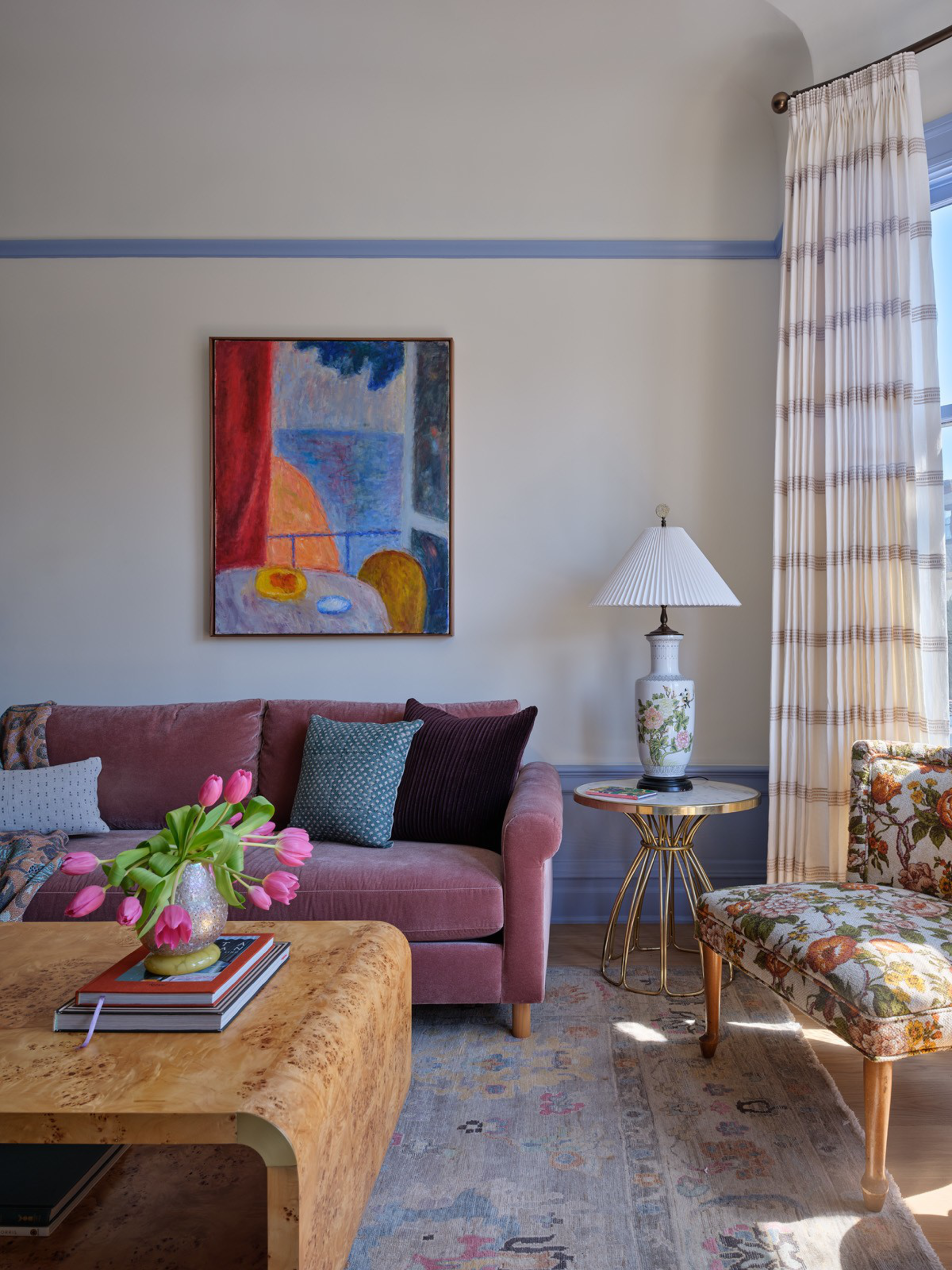 A cozy living room with a mauve sofa, colorful pillows, a floral armchair, a wooden coffee table with tulips, and a side table with a floral lamp.