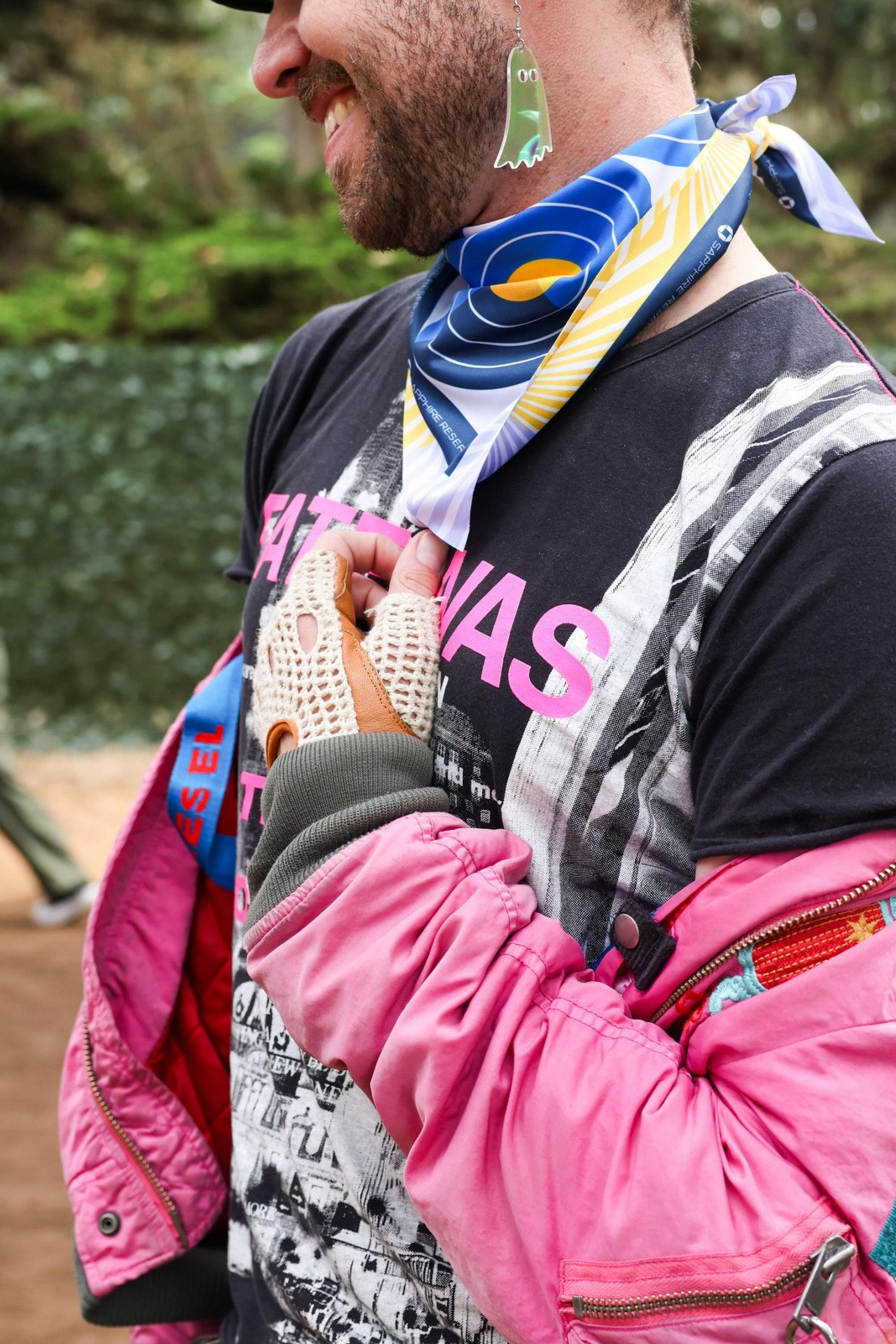 A person wears a pink jacket, black graphic tee, striped pants, a colorful scarf, and an earring shaped like a small ghost. They are smiling, partially obscured from view.