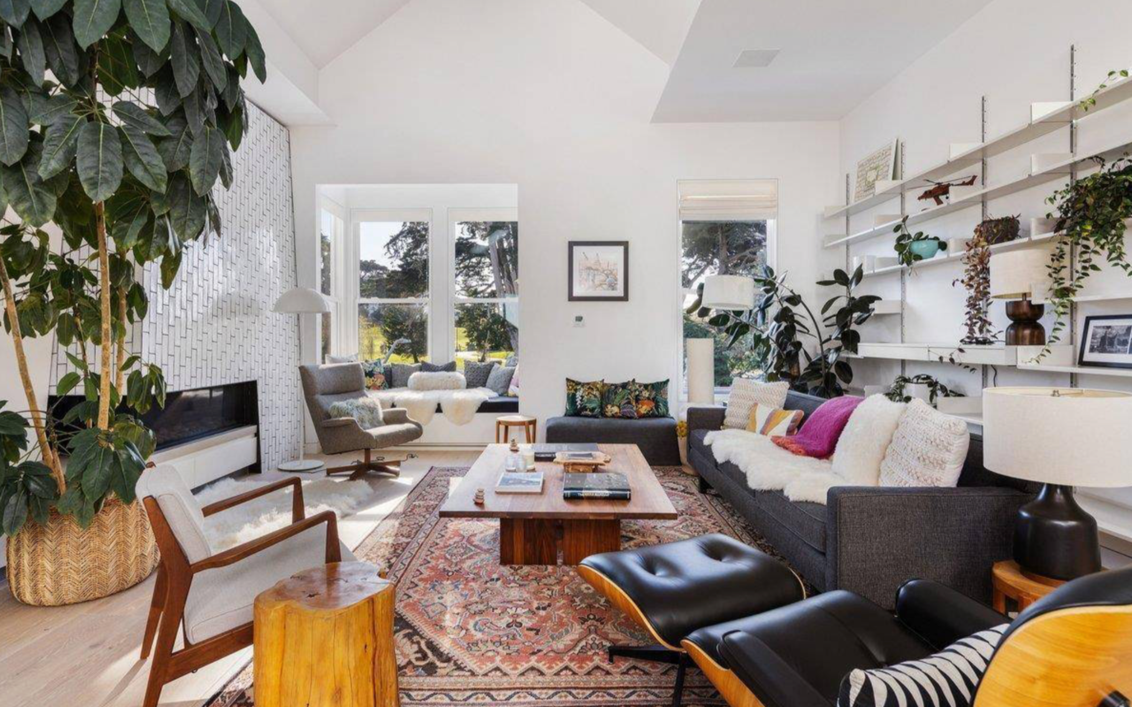 A bright living room features a large area rug, modern gray sofas with colorful pillows, leather lounge chair, wooden furniture, green plants, and large windows.