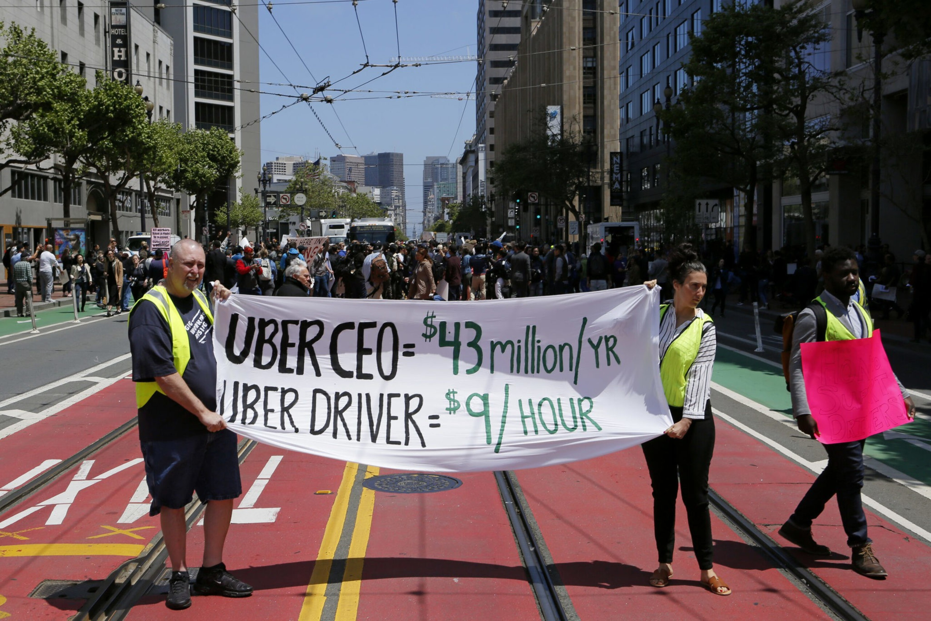 Three protesters stand on a city street holding signs, including one stating the Uber CEO earns $43 million per year and Uber drivers earn $9 per hour.