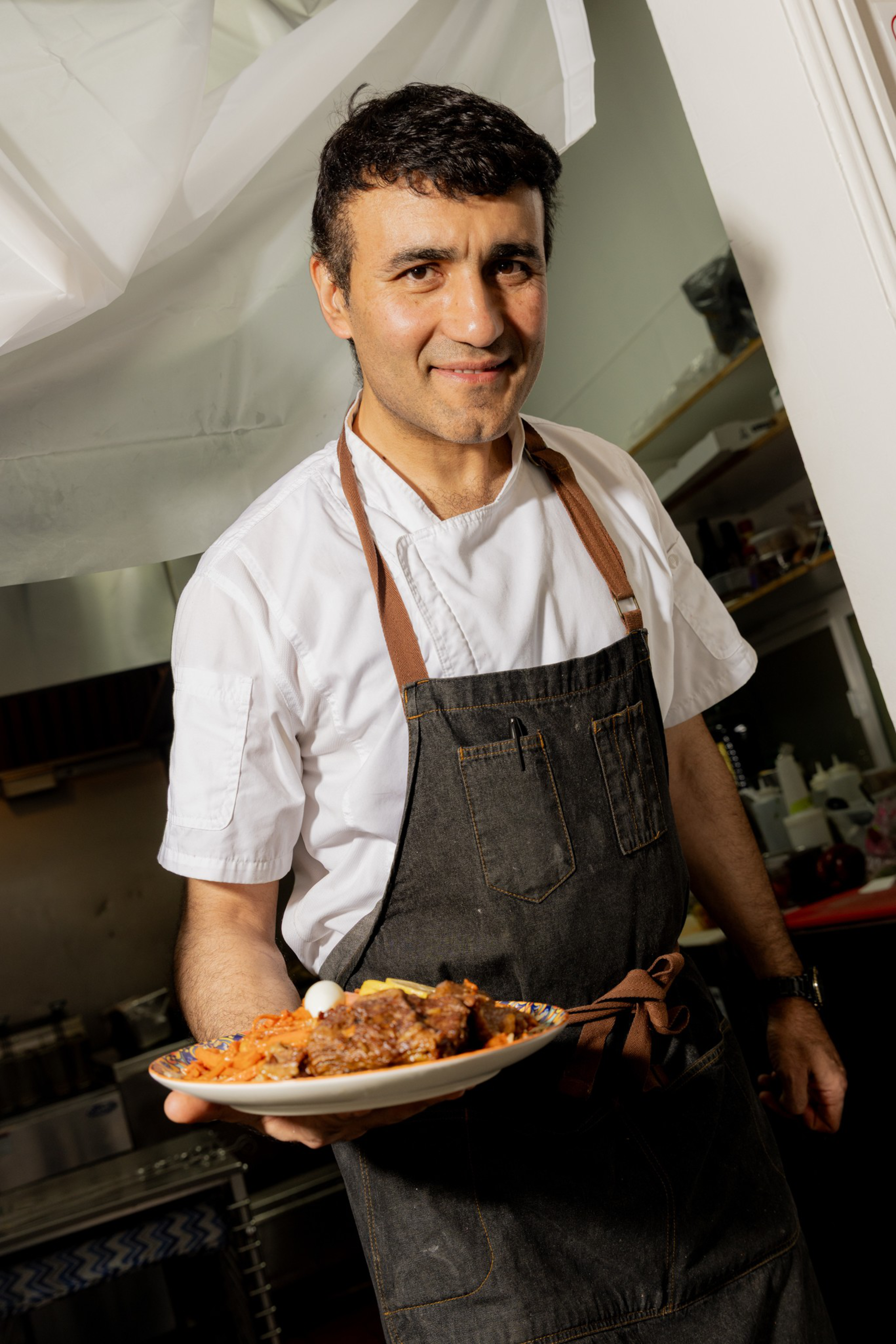 A man in a white chef’s jacket and dark apron is holding a plate of food, smiling, standing in a kitchen.