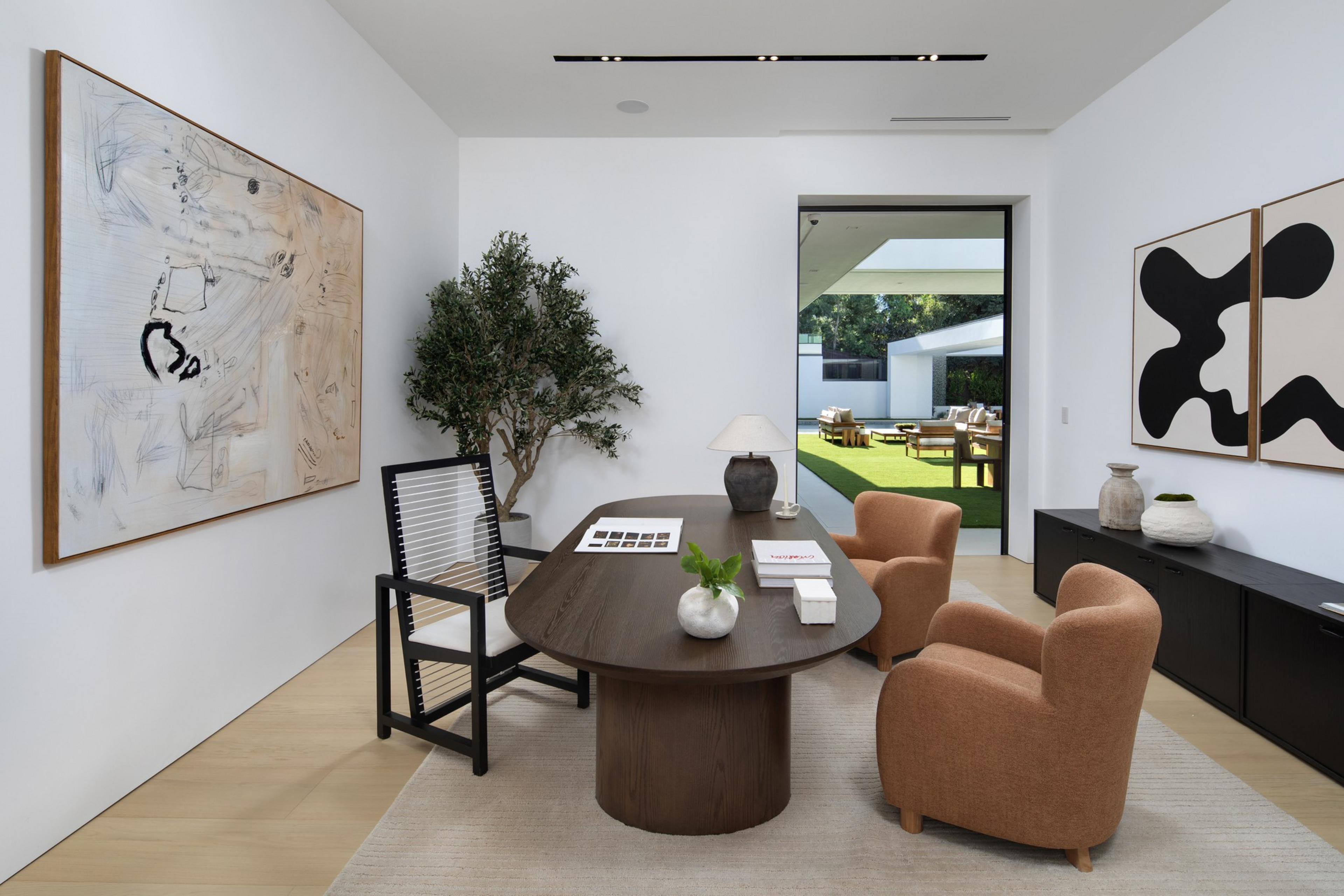 A modern office features a dark wood oval table, a black chair with white straps, two orange armchairs, abstract art, and a view of an outdoor patio with green grass.