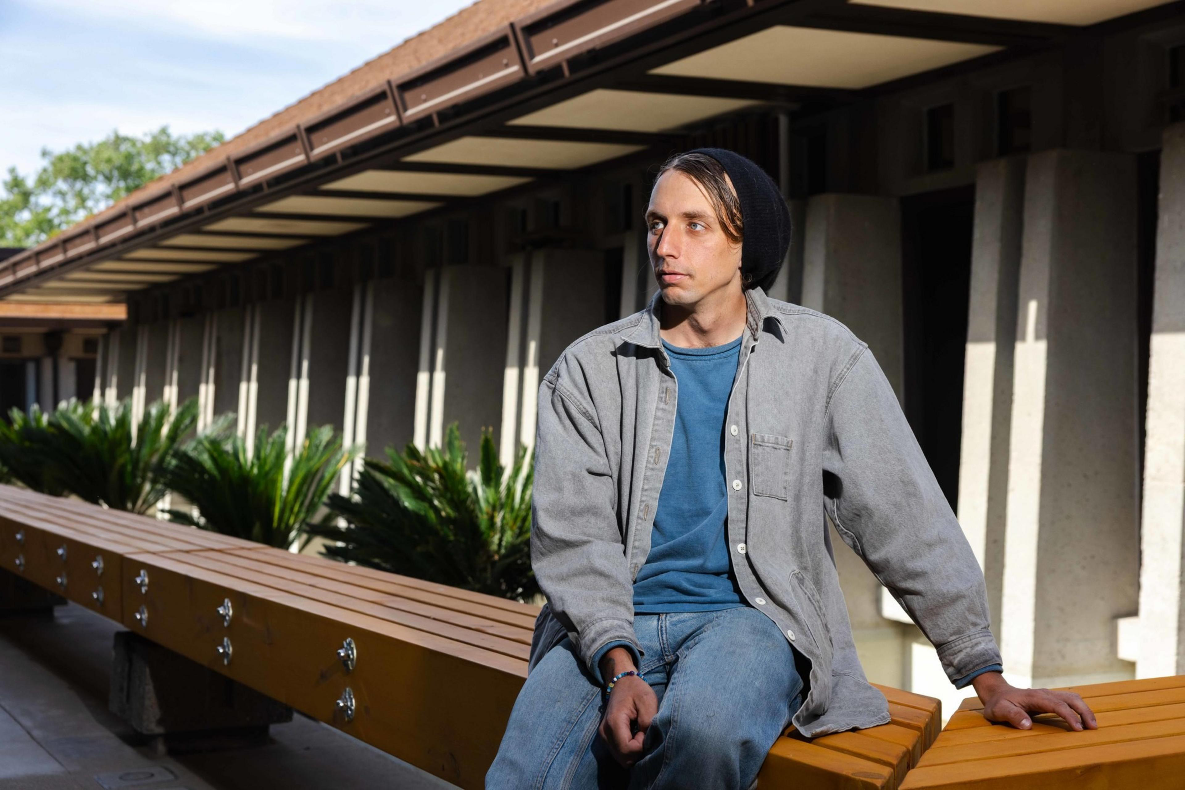 A person wearing a beanie, blue shirt, and gray overshirt sits on a yellow bench outside a building with white columns and plants.