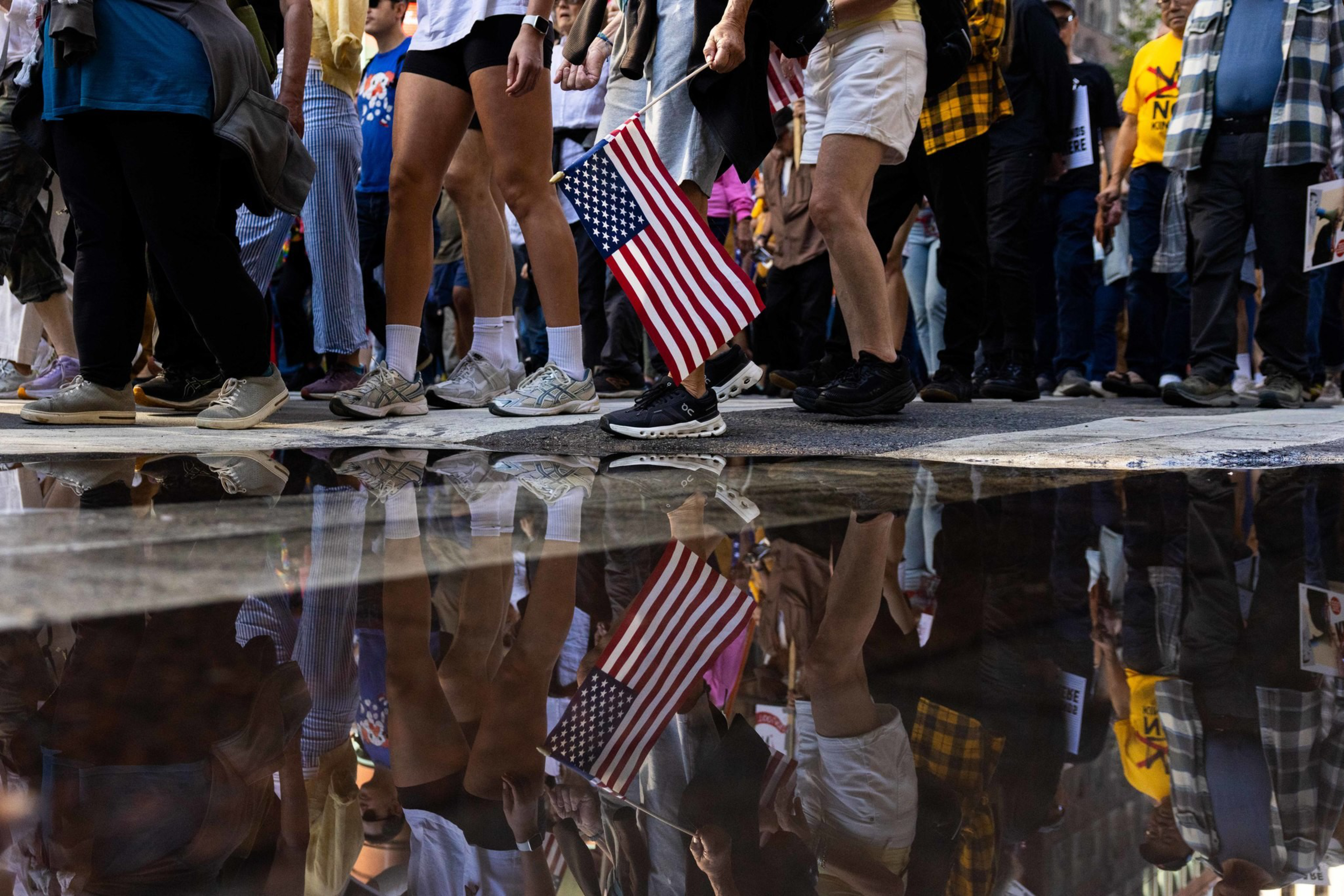 A group of people walk closely together, one holding an American flag, with their legs and the flag reflected clearly in a water puddle below.