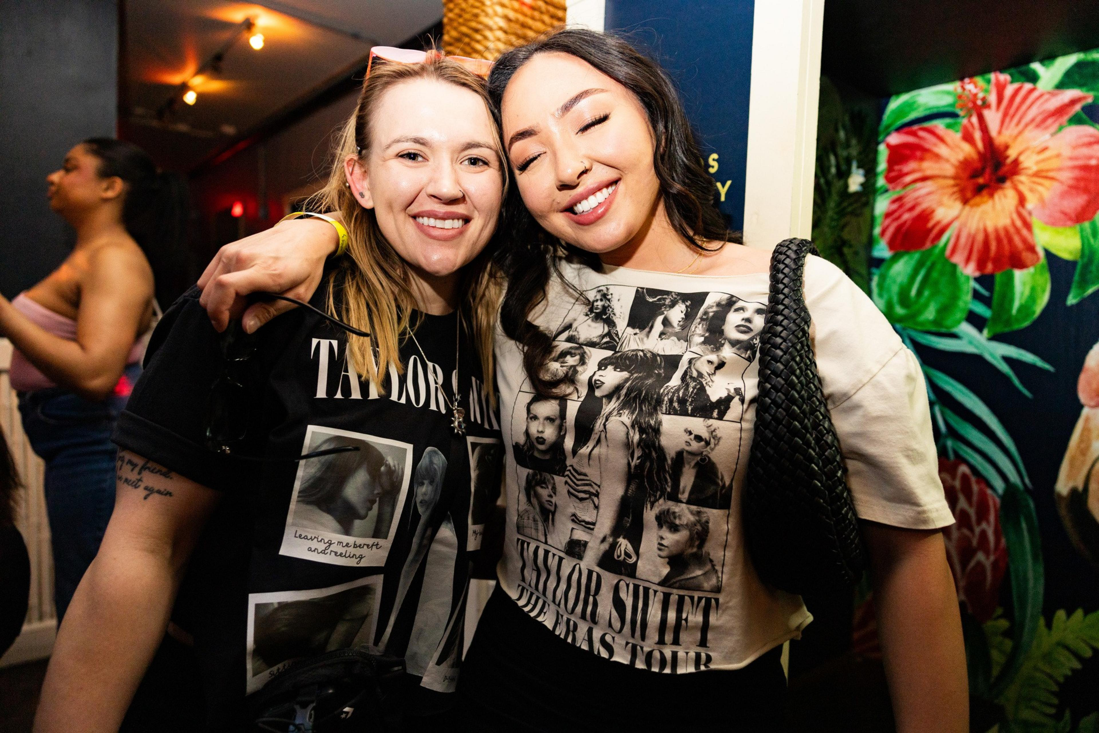 Two smiling women in Taylor Swift T-shirts pose together in a room with floral wallpaper.