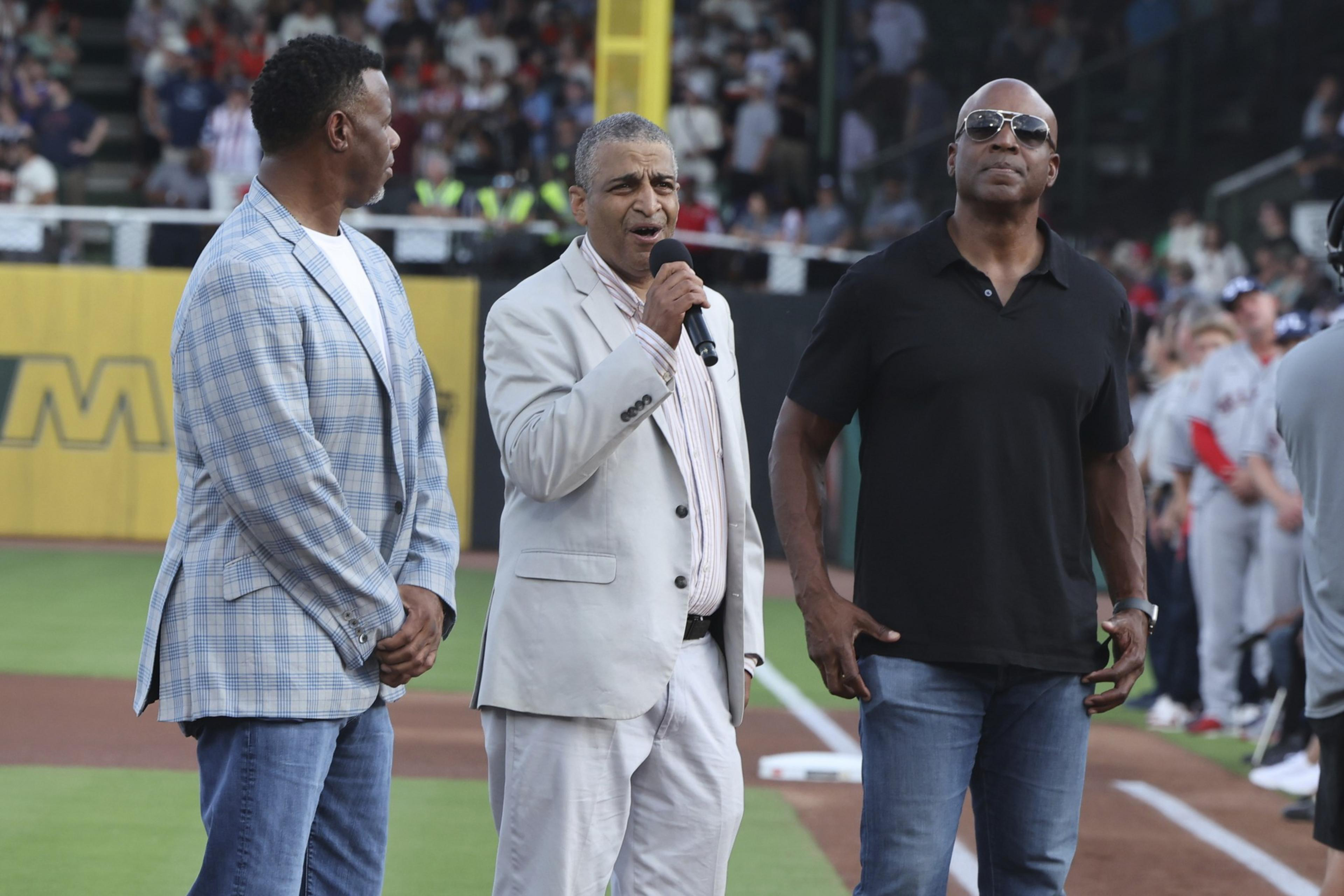Three men stand on a baseball field. One wears a plaid blazer, another a light suit singing into a microphone, and the third in a black shirt and sunglasses.