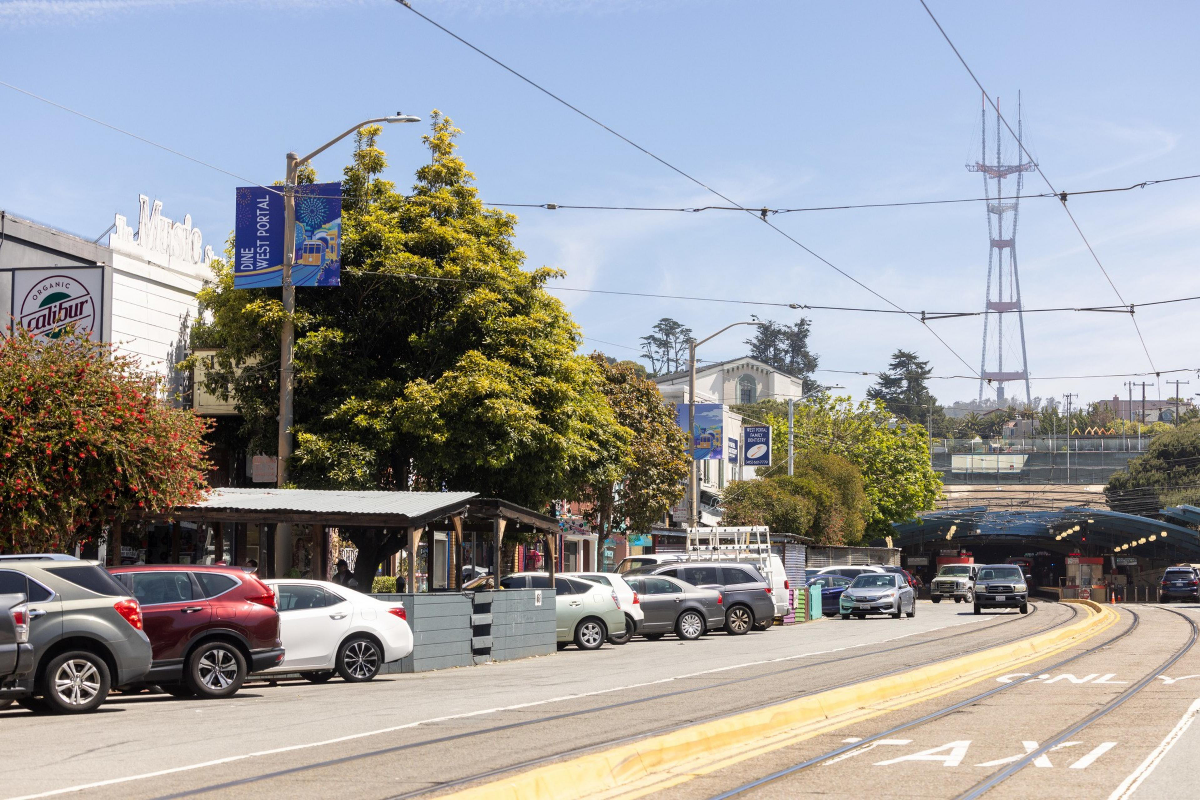 The image shows a street with parked cars along the side, tree-lined sidewalks, and a visible cafe. Overhead power lines stretch across the scene, and a tall broadcast tower can be seen in the background.