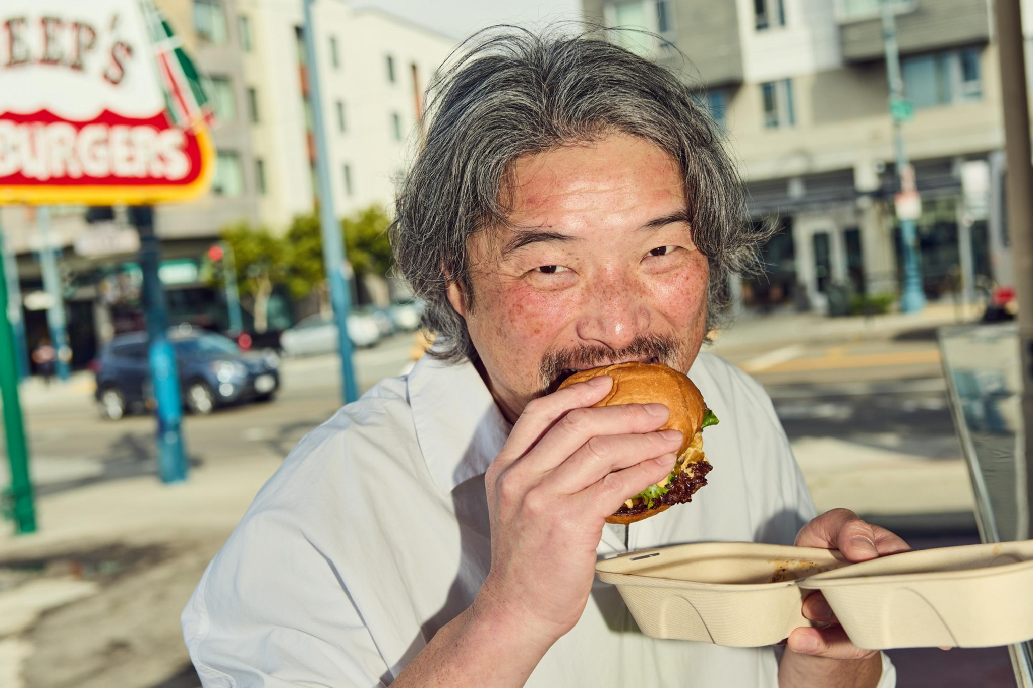 A man with grey hair is enjoying a large burger while standing outside, holding a takeout container. A street and buildings are visible in the background.