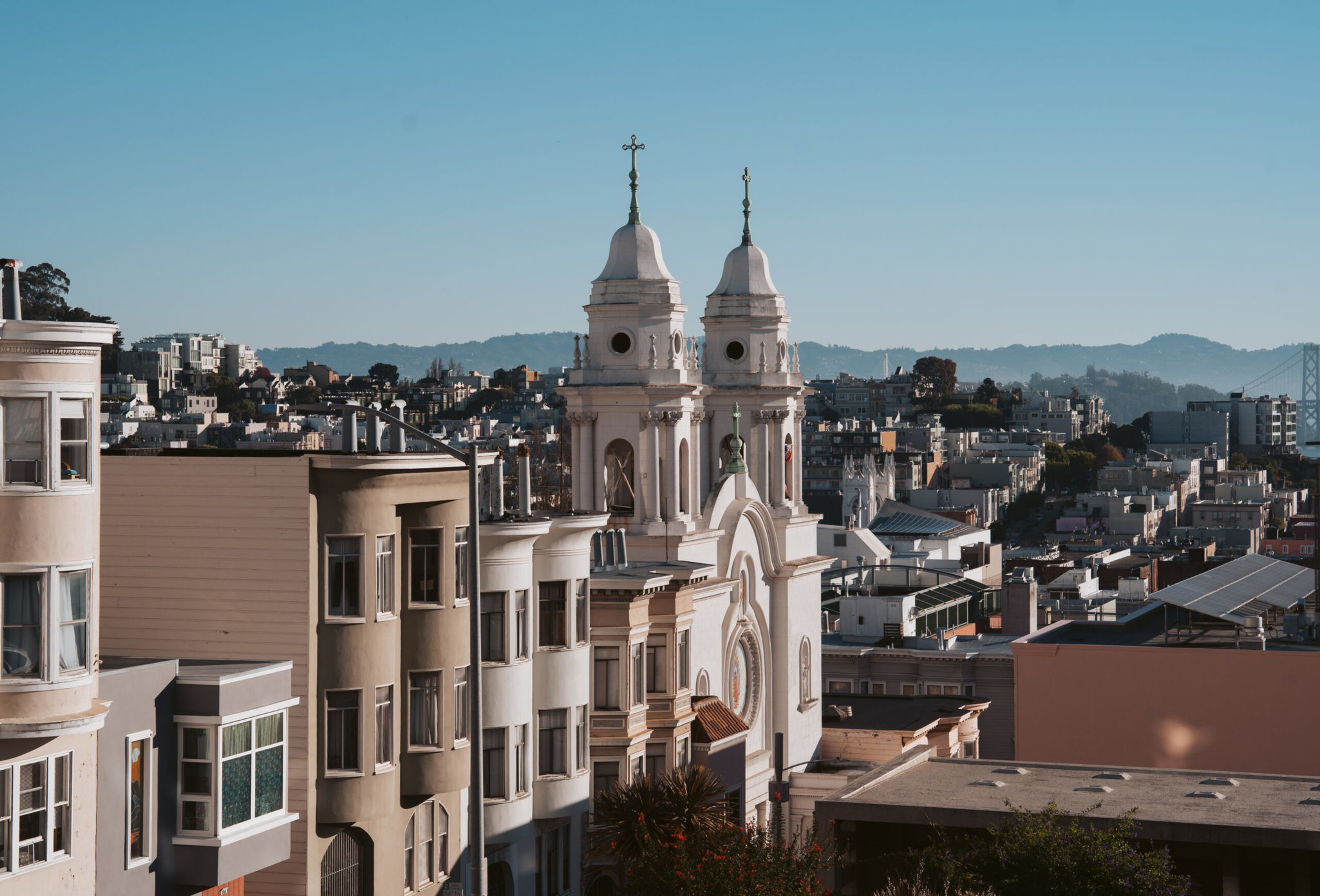 A cityscape featuring a church with two tall, white towers topped with crosses, surrounded by varied residential buildings under a clear blue sky.