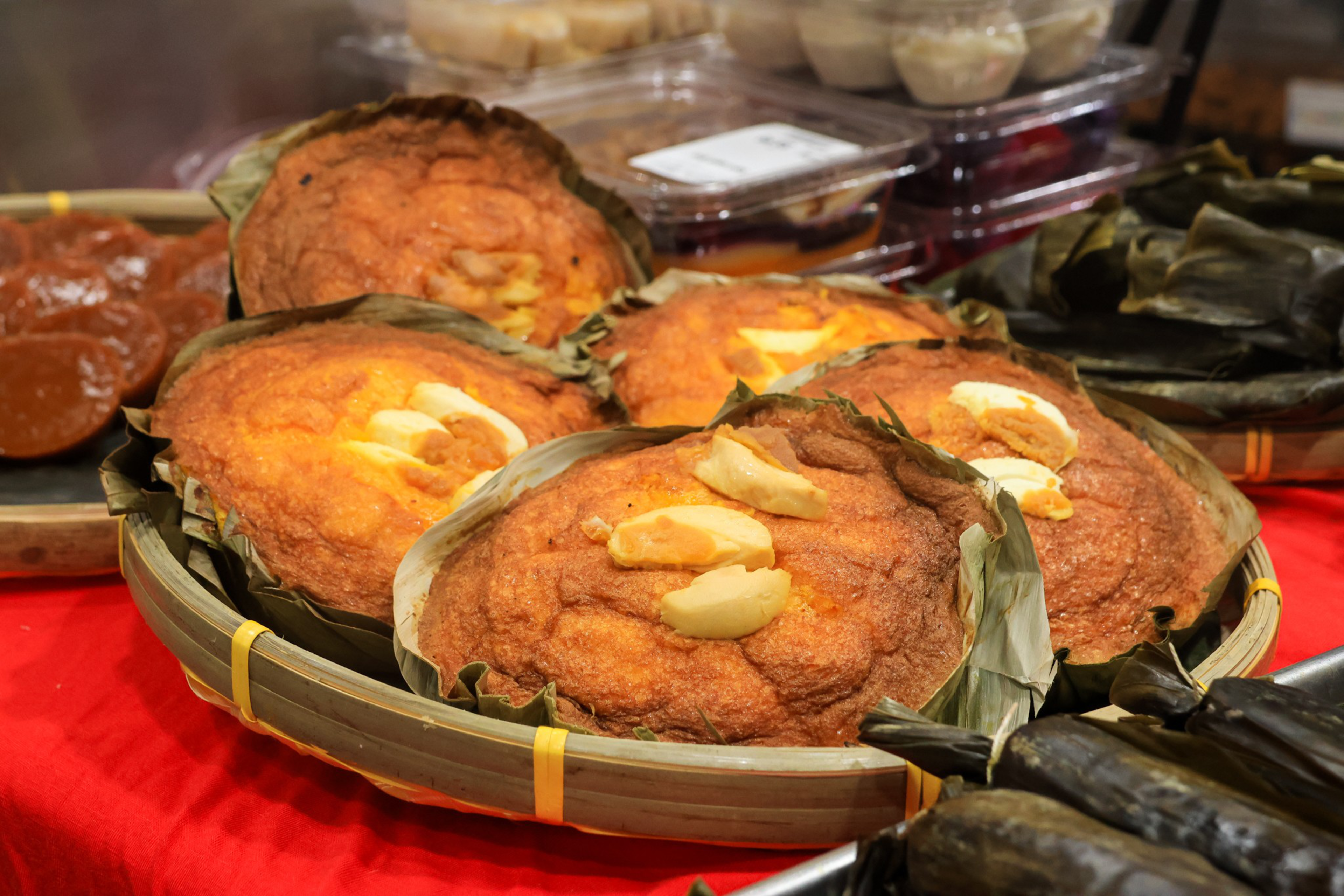 Four round, brownish-orange baked cakes topped with slices of egg are placed in a woven bamboo basket lined with green leaves.
