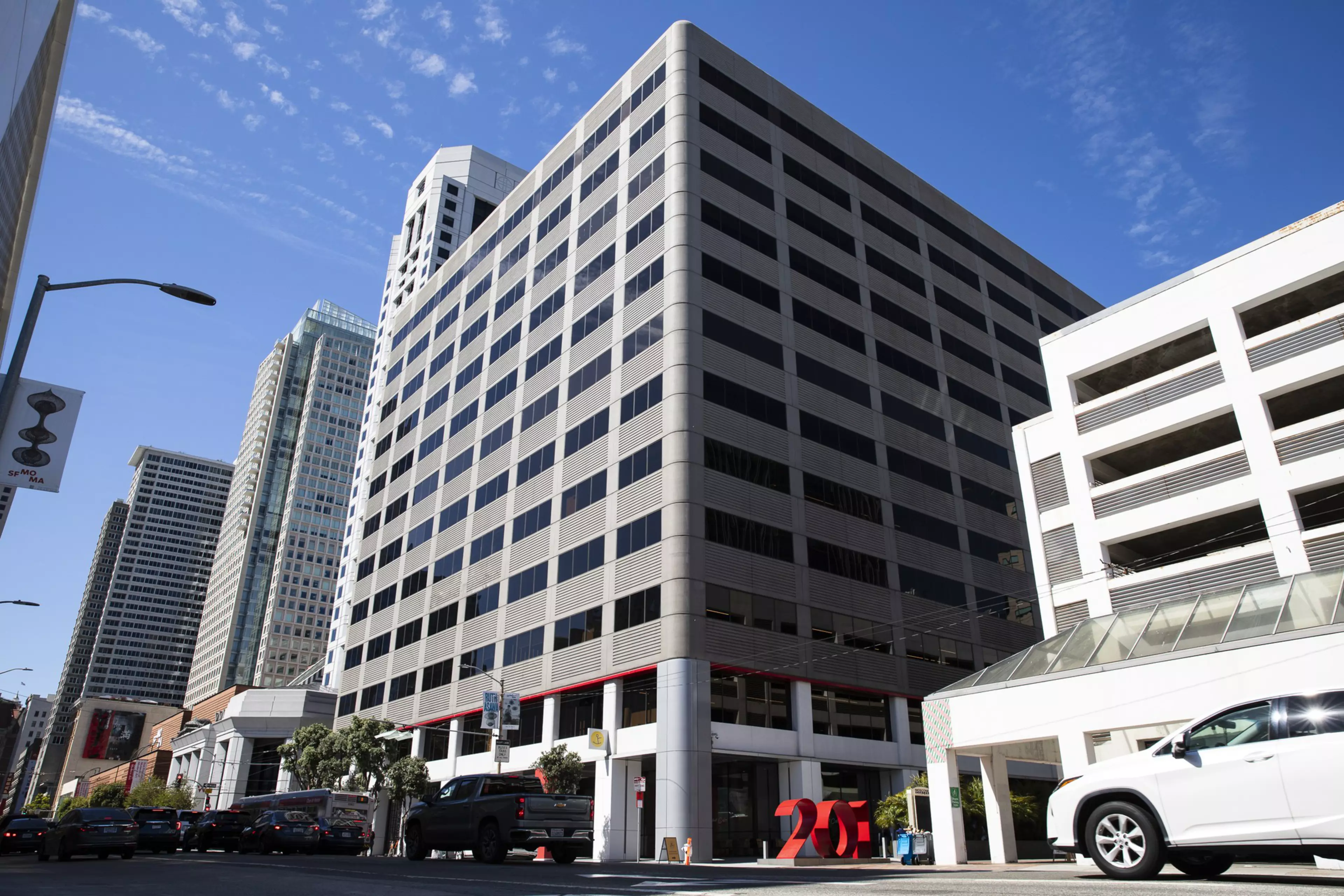 A large modern office building with many windows sits on a city street corner, next to a white multi-level parking garage and several parked vehicles.