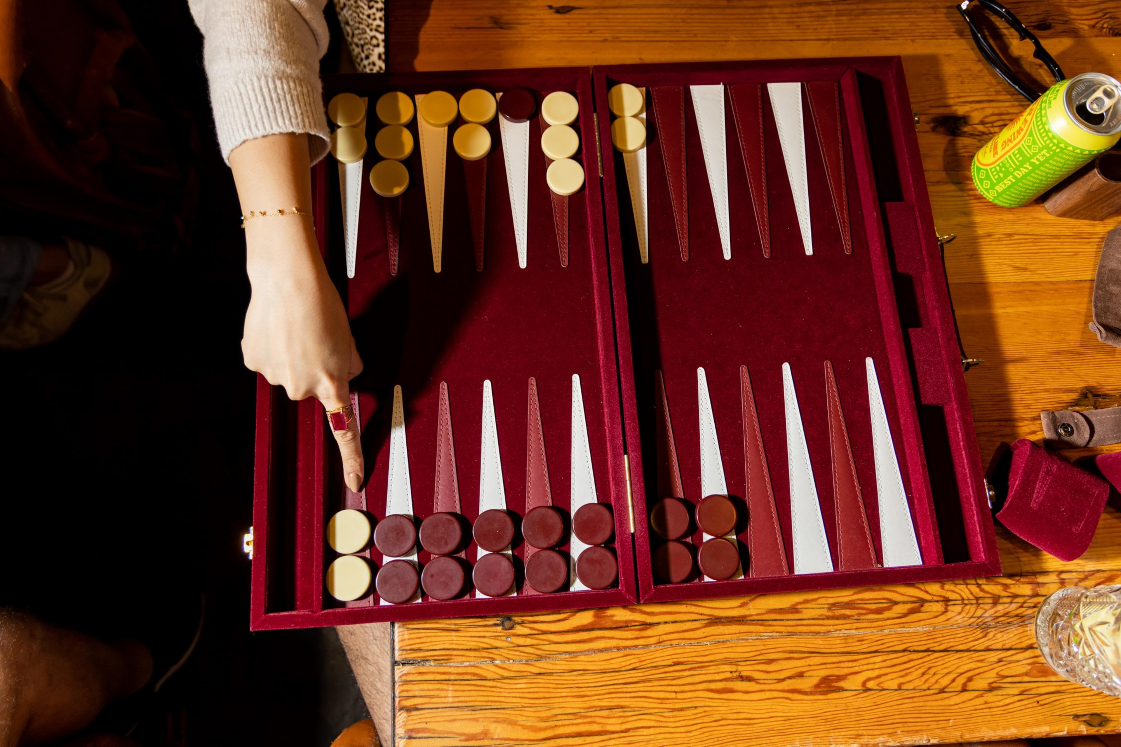 A backgammon board with red and white points, brown and cream pieces in starting positions, and a person’s hand pointing at a brown piece.