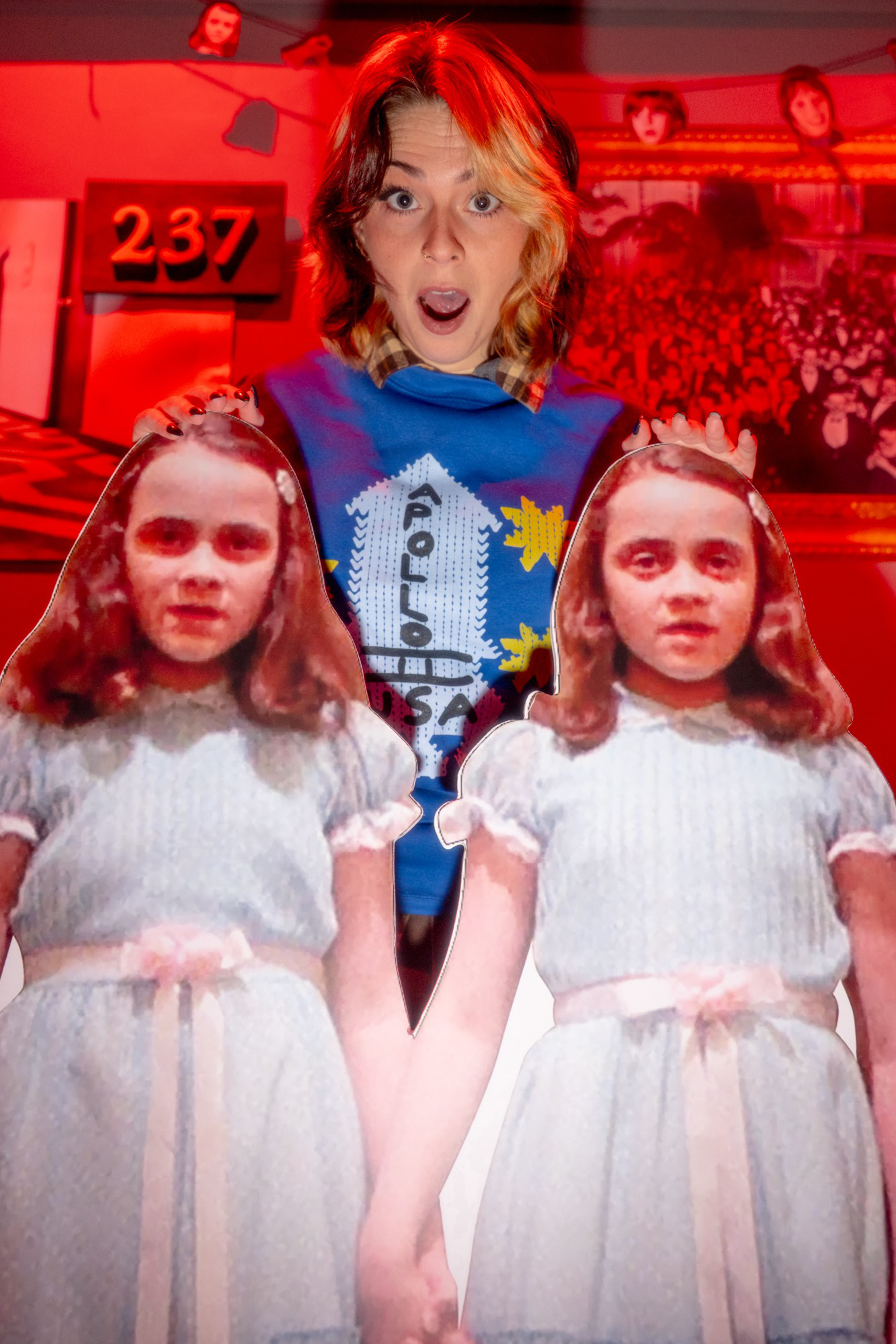 A person with half-red, half-blonde hair looks surprised while holding cutouts of twin girls in white dresses with pink ribbons. The background is red-lit.