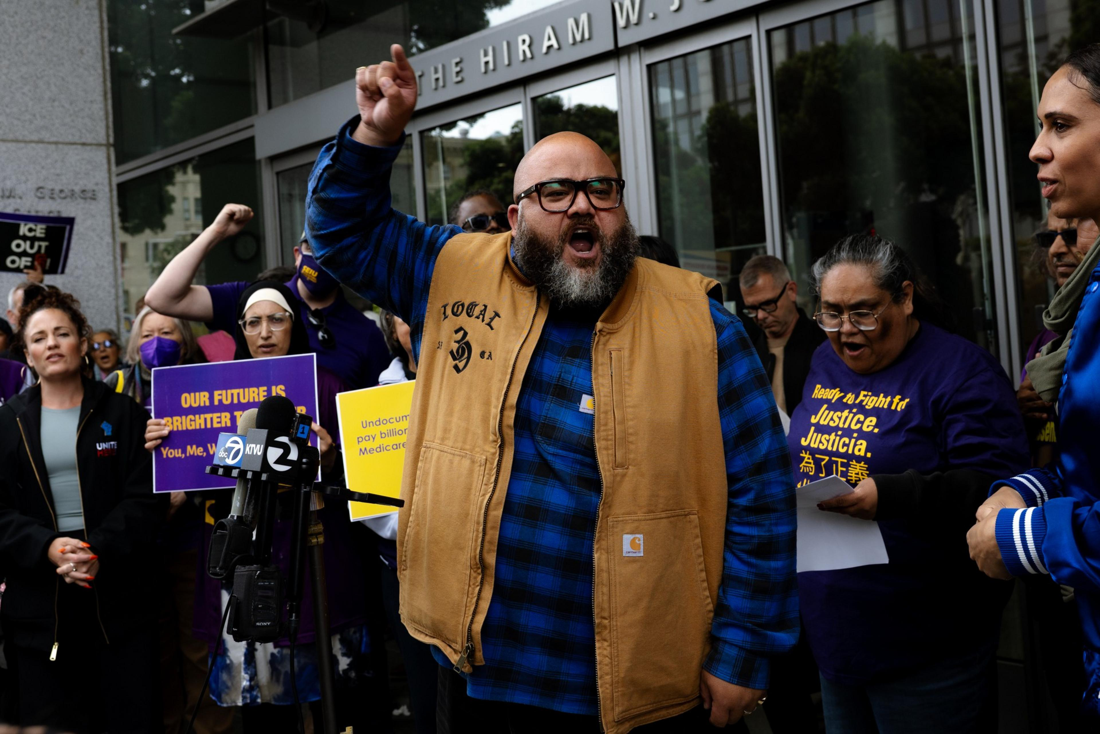 A group of people are protesting outside a building. A man in a blue plaid shirt and tan vest raises his fist passionately. Others hold signs emphasizing justice.