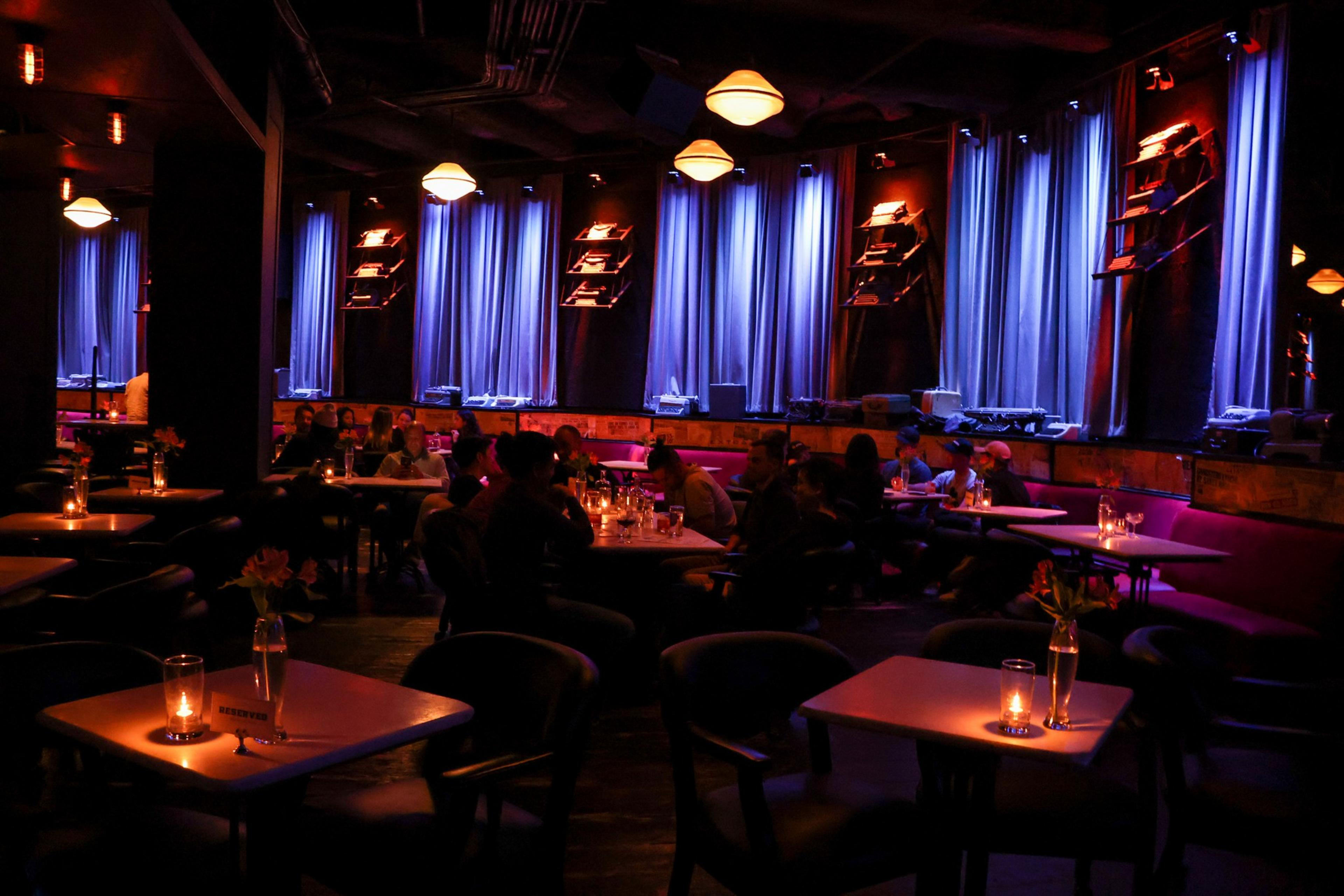 A dimly lit lounge with tables and chairs, featuring candles and flowers. Blue curtains and warm light create a cozy atmosphere. People are seated and chatting.