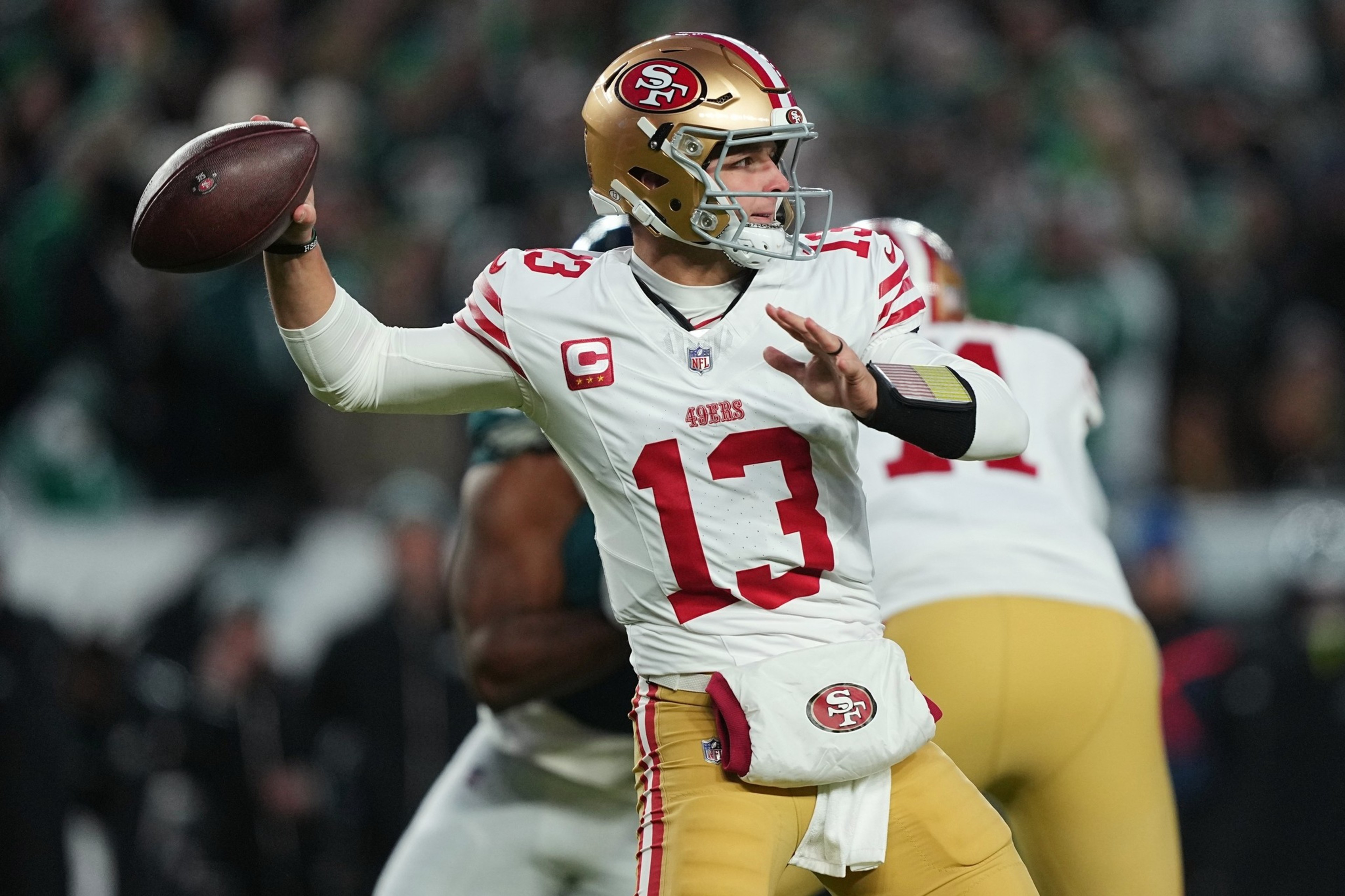 A football player in a white San Francisco 49ers jersey and gold helmet prepares to throw a pass during a game.
