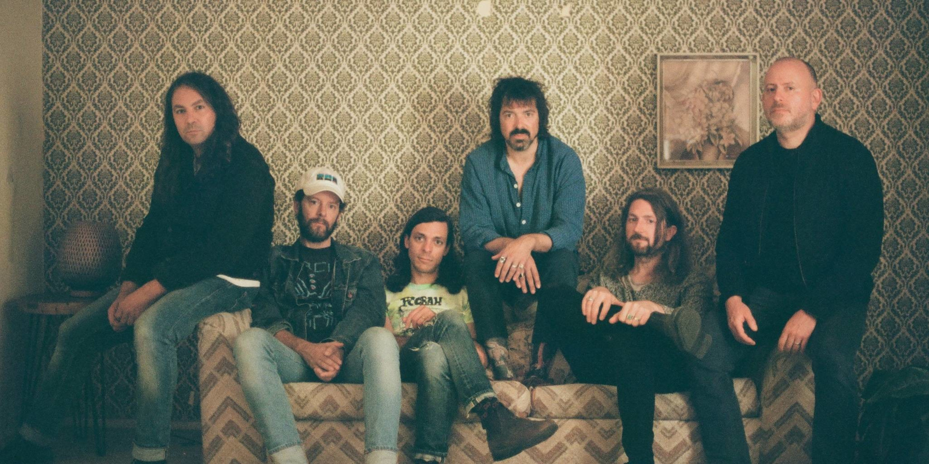 Six men with casual attire and varied hairstyles sit and stand around a patterned couch against a vintage wallpapered wall with a framed picture.