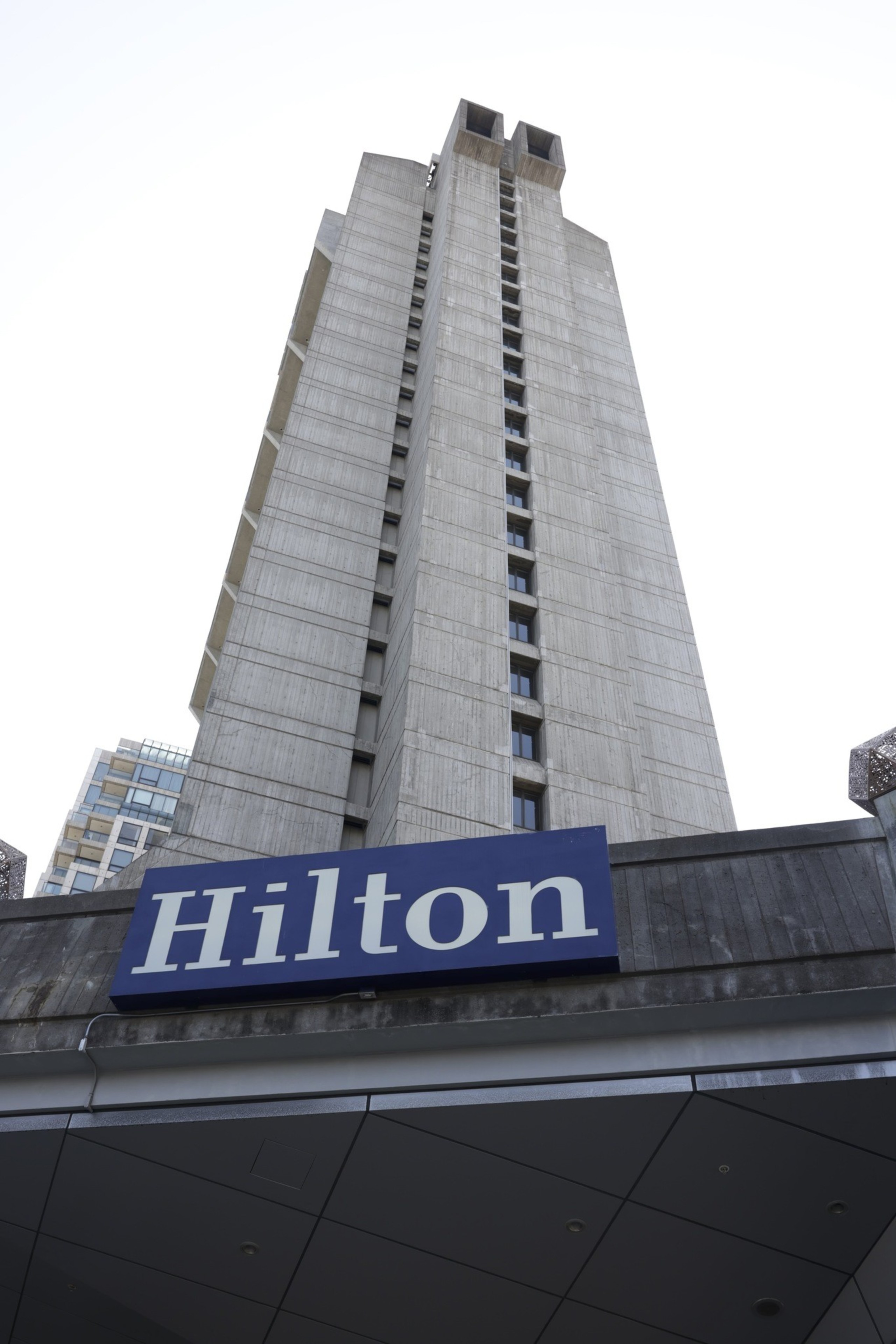 The image shows a tall, modern concrete building viewed from below, with a blue sign reading &quot;Hilton&quot; prominently displayed at the bottom.