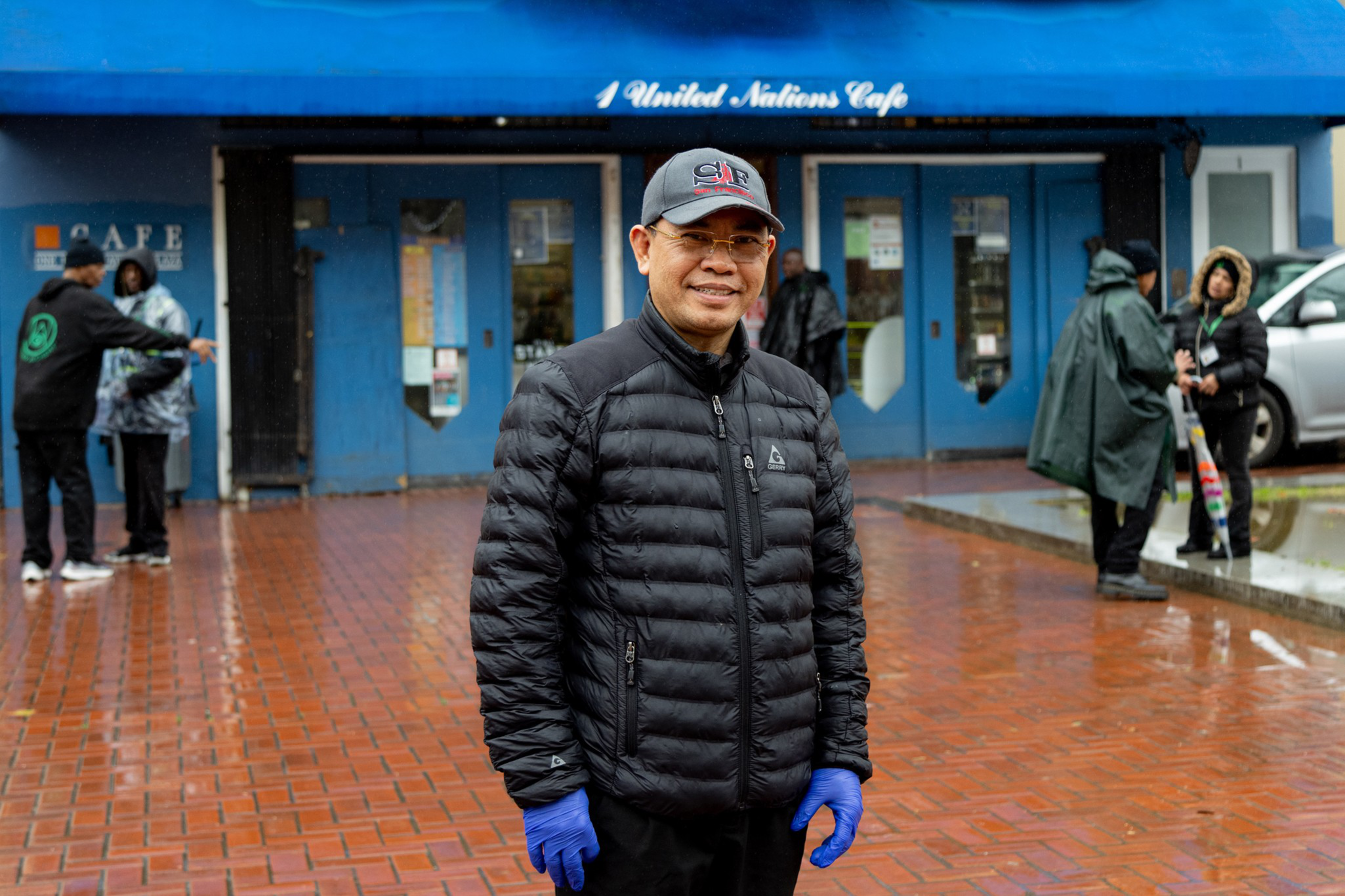 A man in a black jacket and blue gloves stands smiling on a wet brick street; blurry people by a blue cafe in the background.