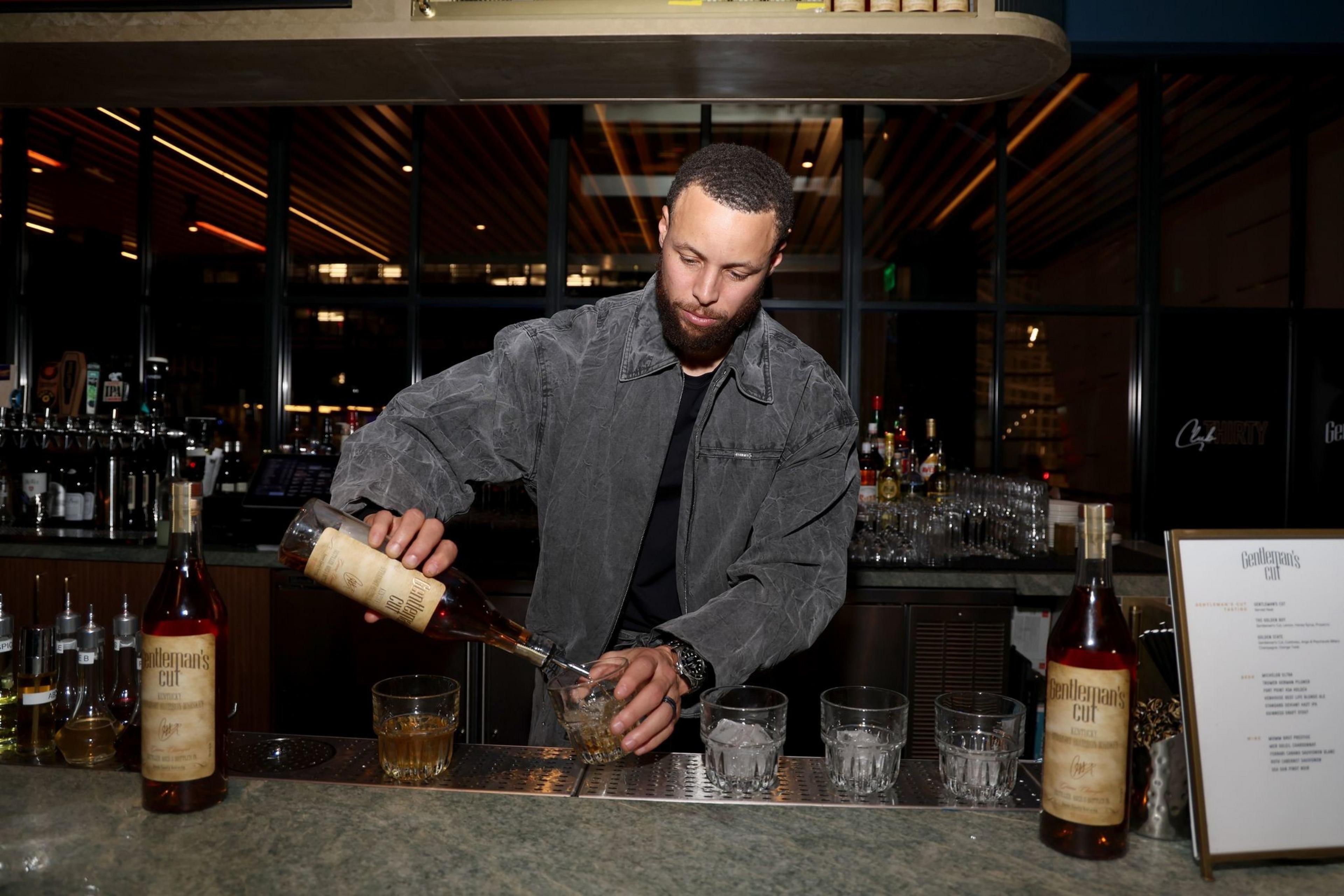 A man with a beard pours whiskey from a bottle into a glass at a bar counter with several other glasses and bottles.