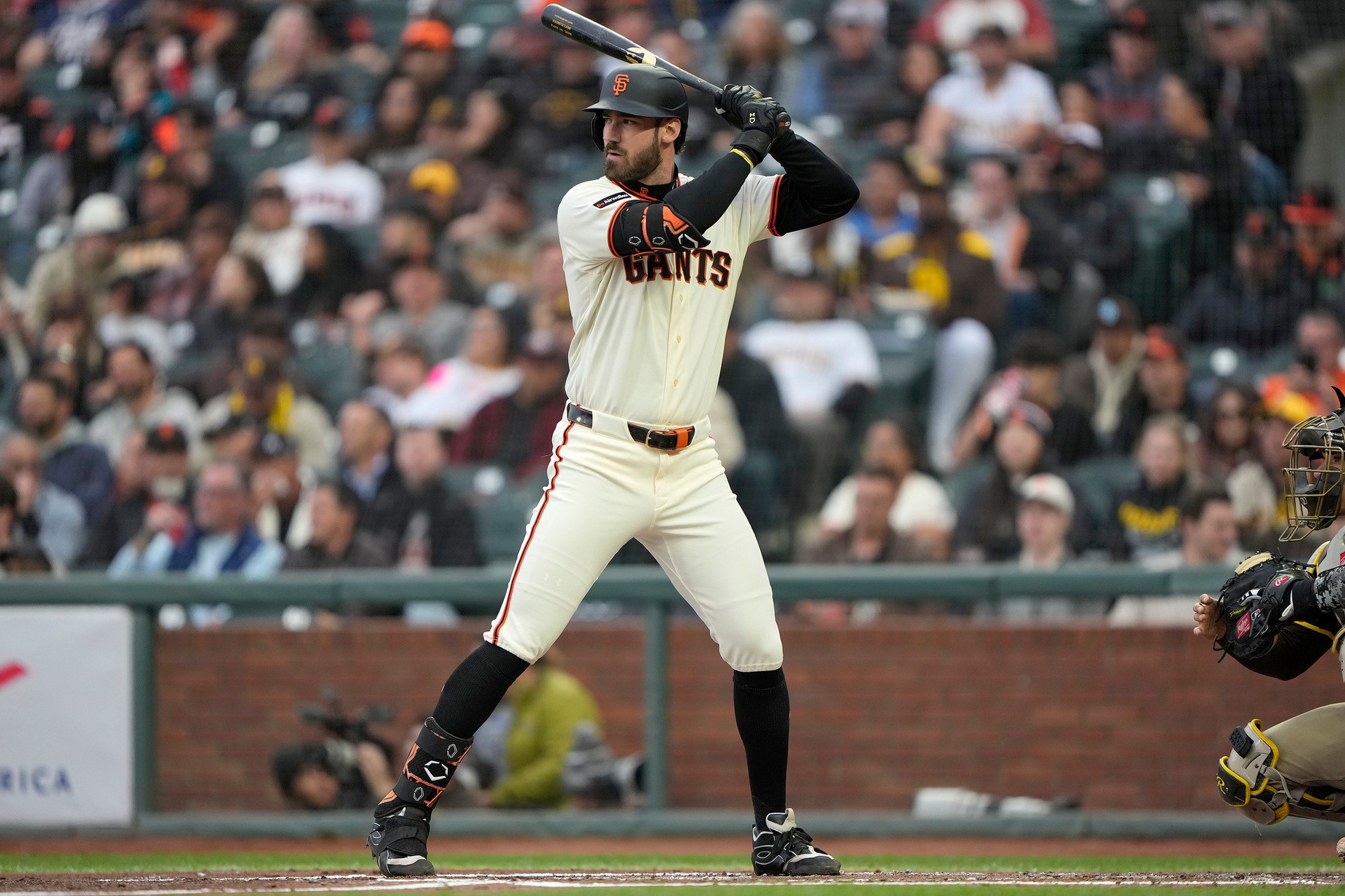 A baseball player in a San Francisco Giants uniform stands ready to bat, with a catcher crouched behind home plate and a crowd in the background.