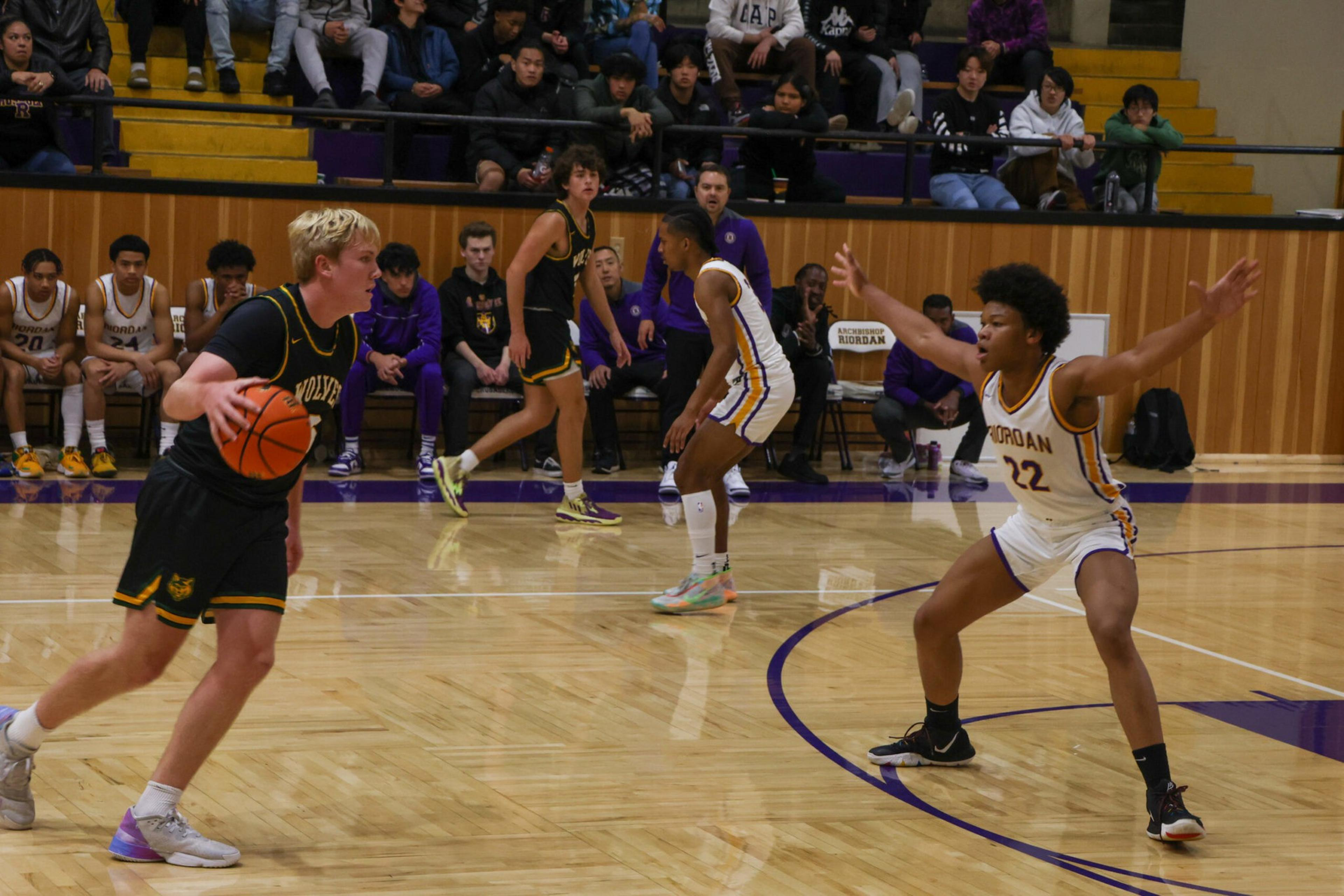 A basketball player in a black uniform dribbles the ball while being guarded by two players in white uniforms on a gym court with spectators in the background.