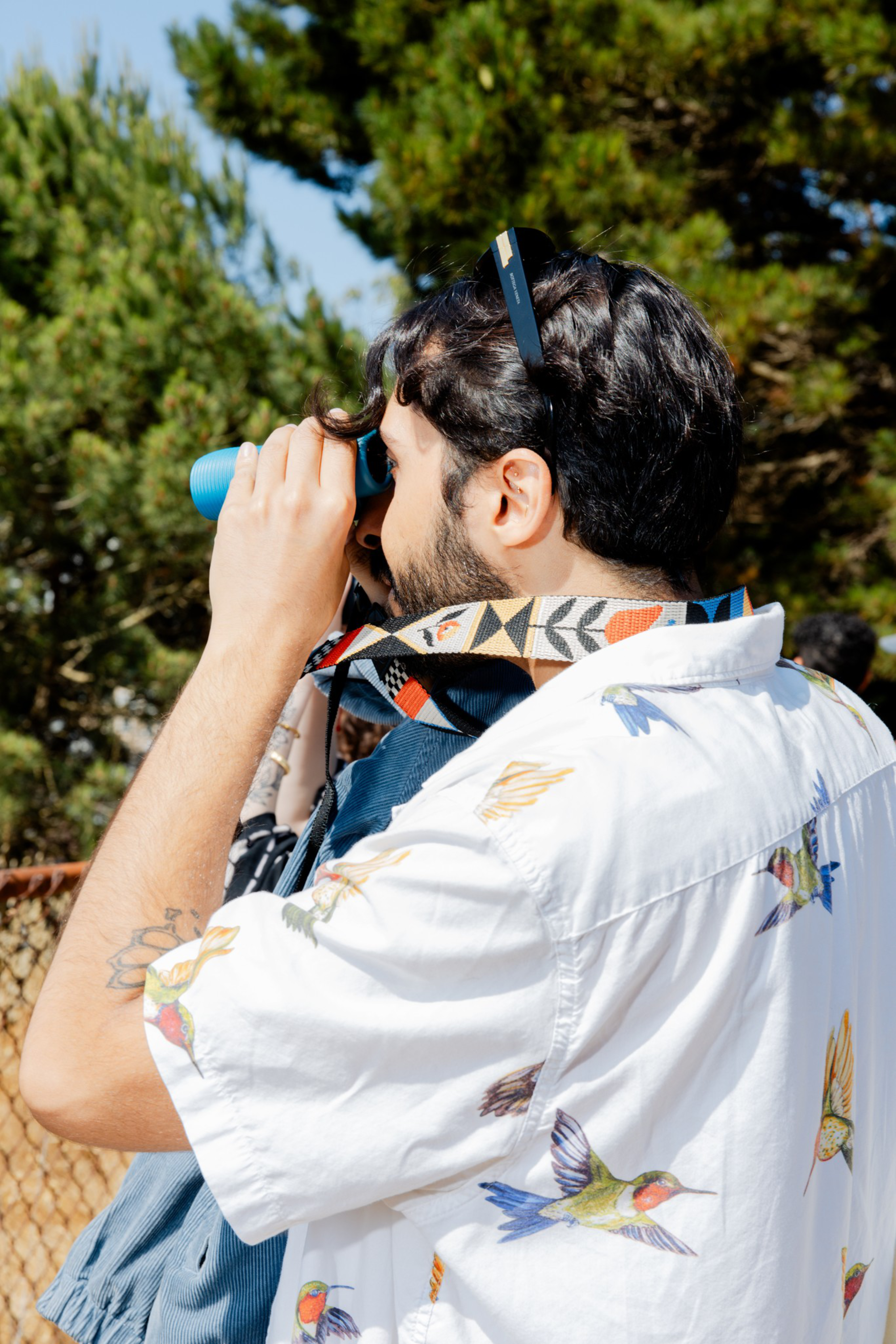 A person in a bird-patterned shirt uses blue binoculars, standing in front of trees, with sunglasses on their head and a camera strap around their neck.
