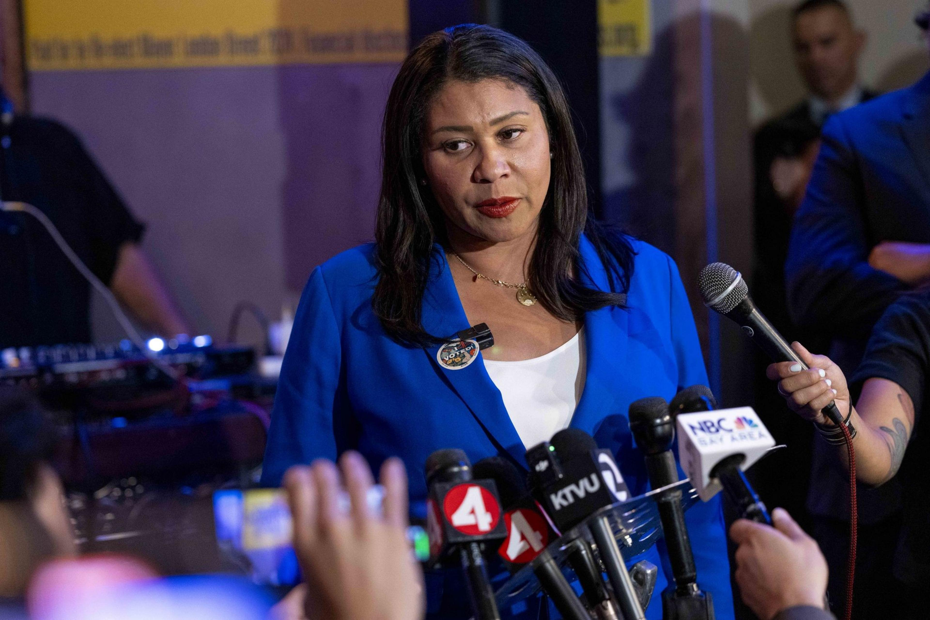 A woman in a blue blazer stands before a cluster of microphones from various news outlets, addressing the media with a serious expression.