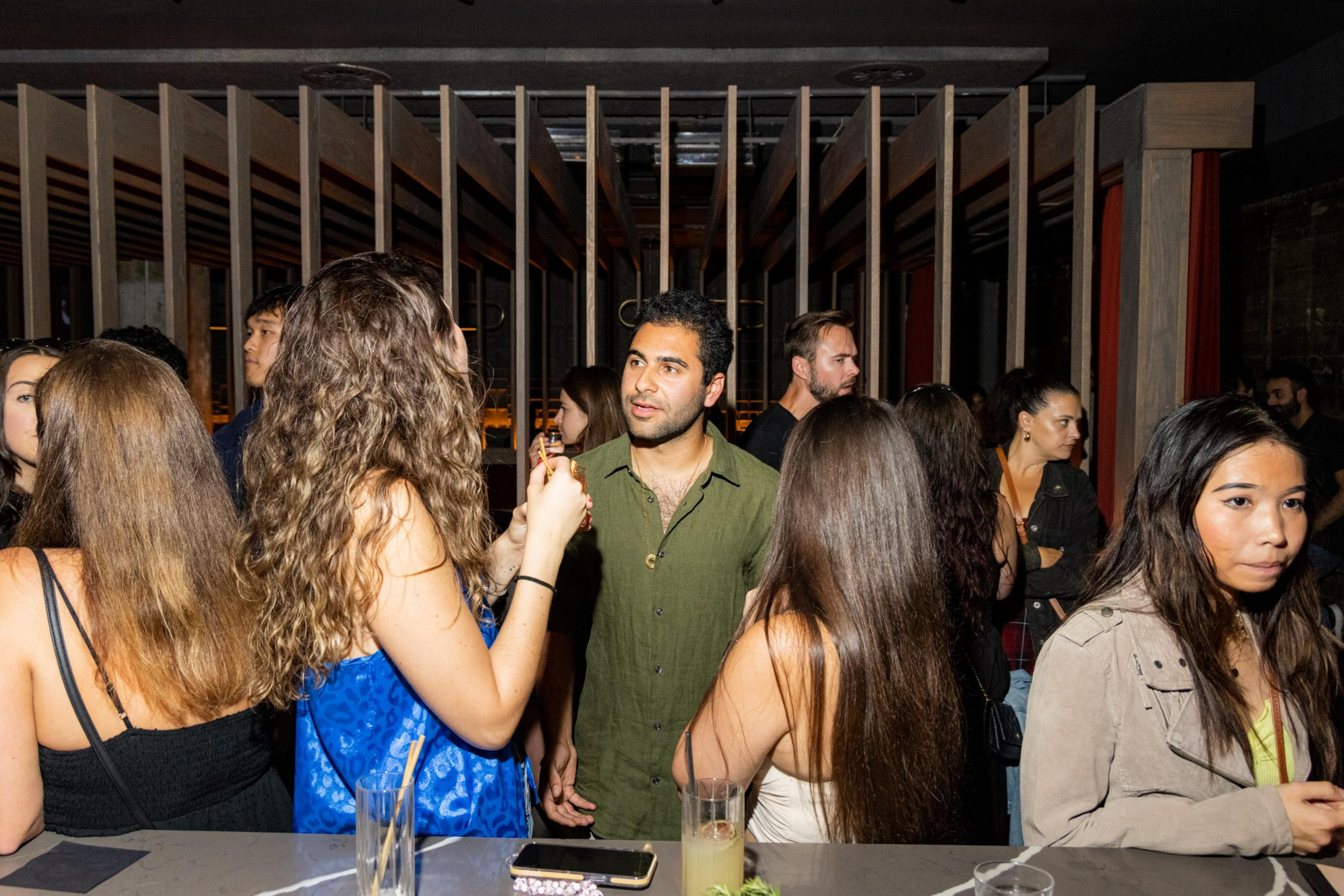 A group of people are socializing at a bar, with drinks in hand and engaging in conversation. The background features wooden accents and dim lighting.