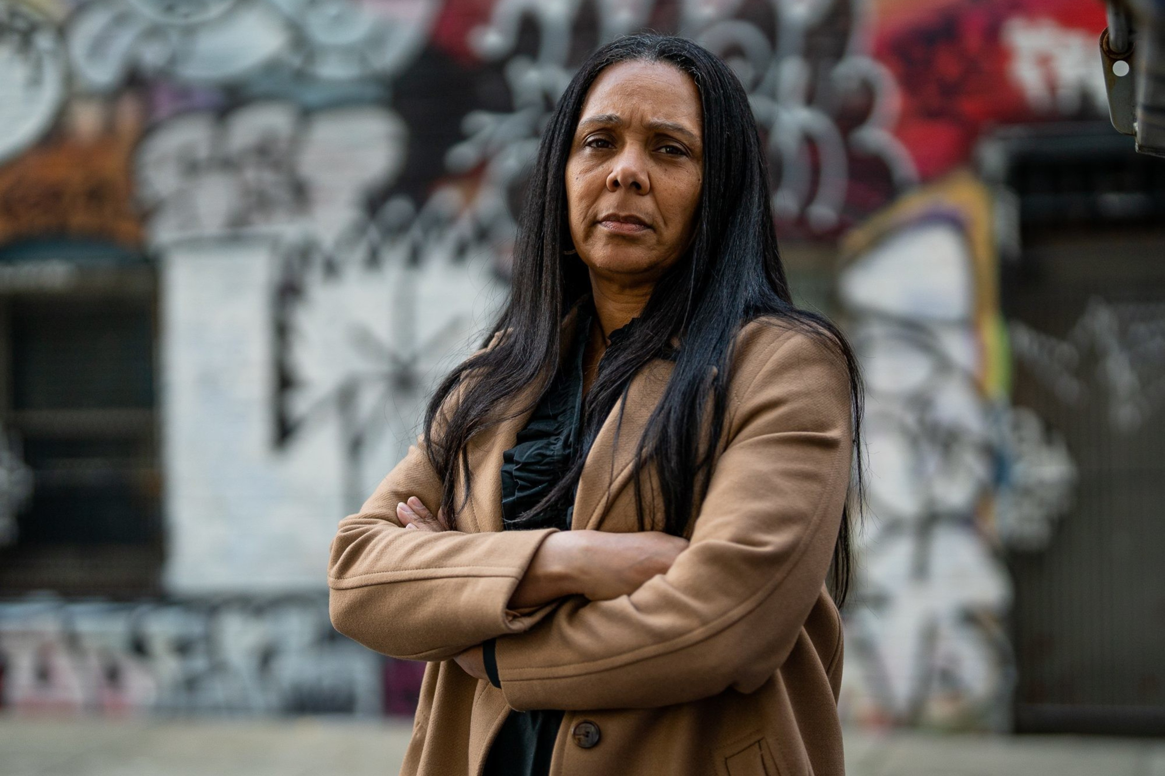 A woman with long dark hair, wearing a tan coat, stands with arms crossed in front of a graffiti-covered wall on an urban street. She has a serious expression.
