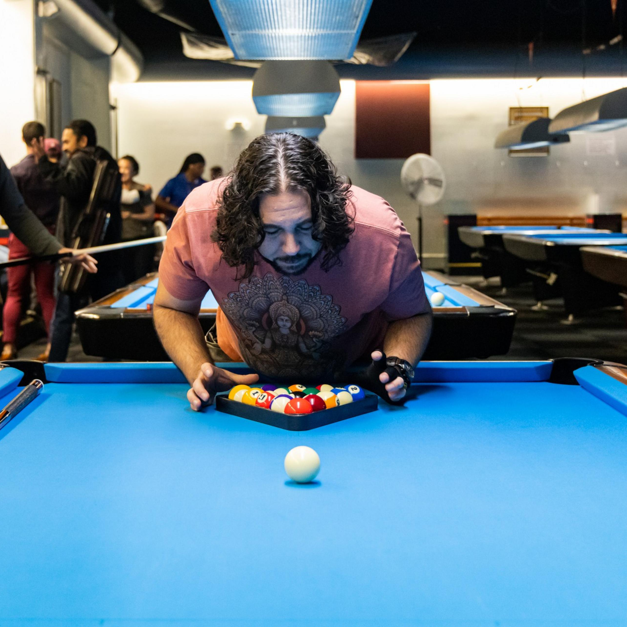 A man with long hair is arranging pool balls on a blue-felted pool table, focusing intently. Several people are in the background, holding pool cues.